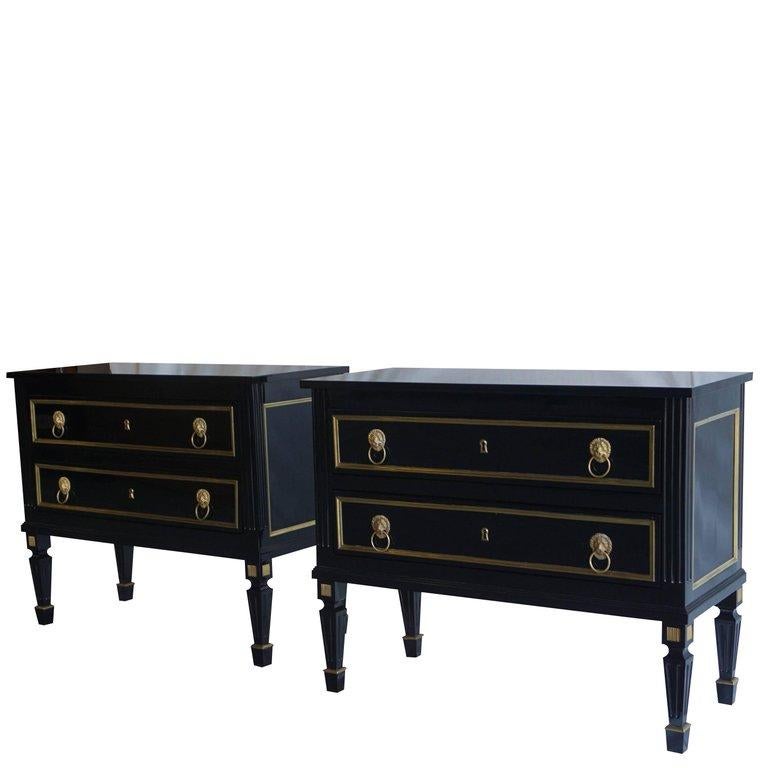 An antique French pair of ebonized wood chests of two drawers on tall tapered legs with brass sabots. The drawers are ornately designed with brass decor and brass lion head hardware, in good condition. The hand carved neoclassical style chests are
