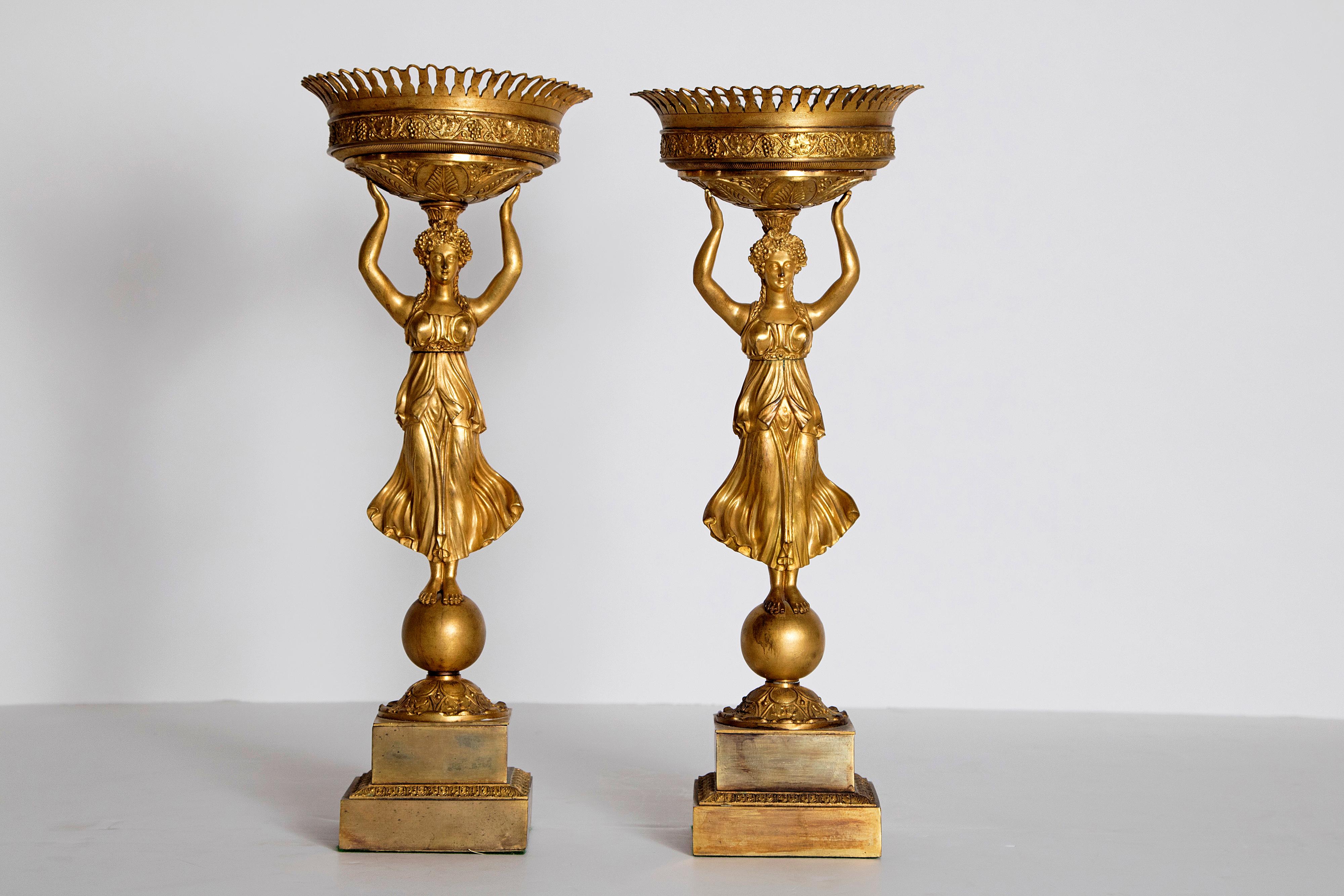 A pair of Empire gilt bronze centerpiece figures of classical women holding a basket aloft with a foliage design and filigree rim. They are standing on an orb above two tiered square bases. The baskets are missing original interior glass liners.