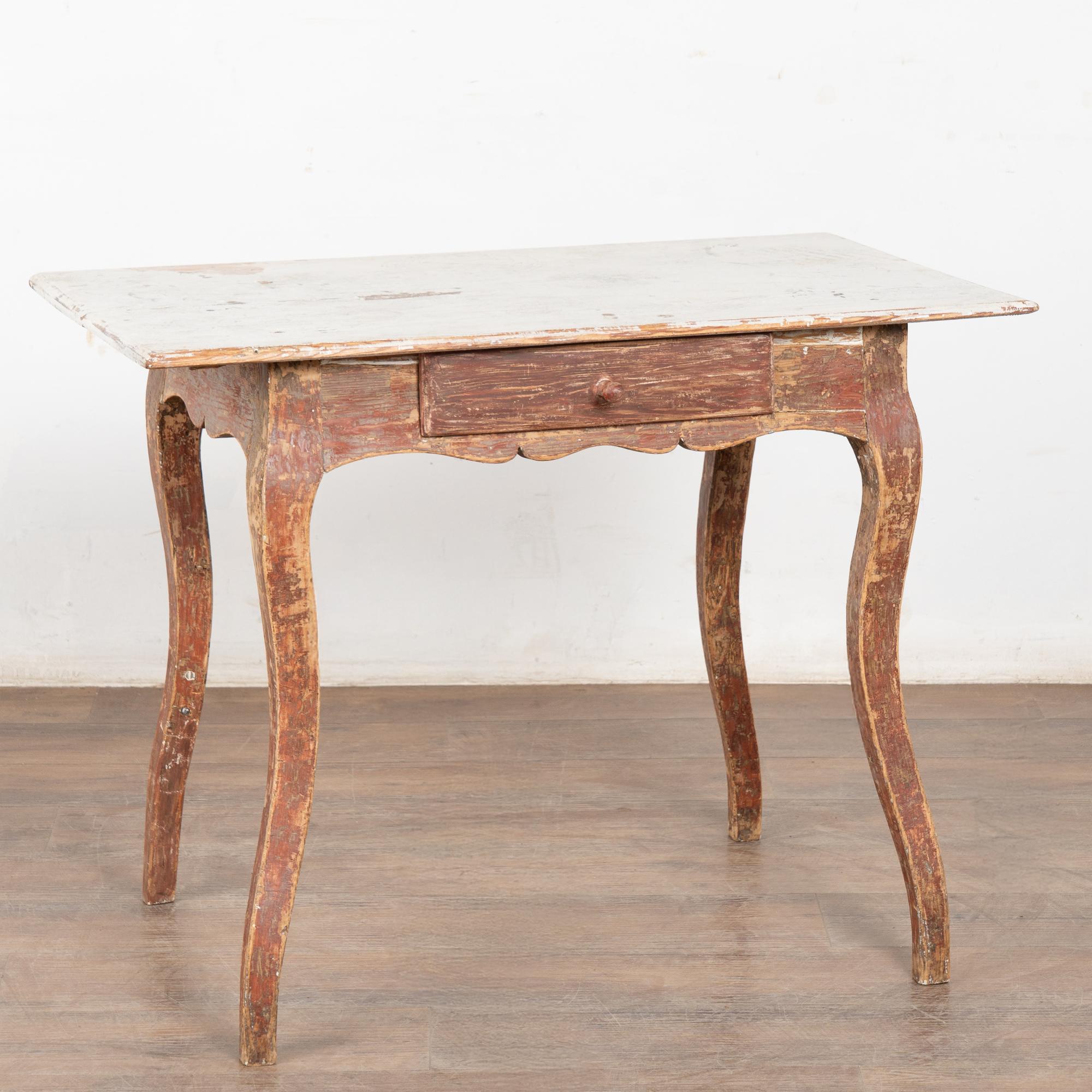The curved cabriole legs add a charming touch to this single drawer pine side table from the Swedish countryside.
The original brick red paint has been scraped and distressed revealing the natural pine below. The contrasting white/gray painted top