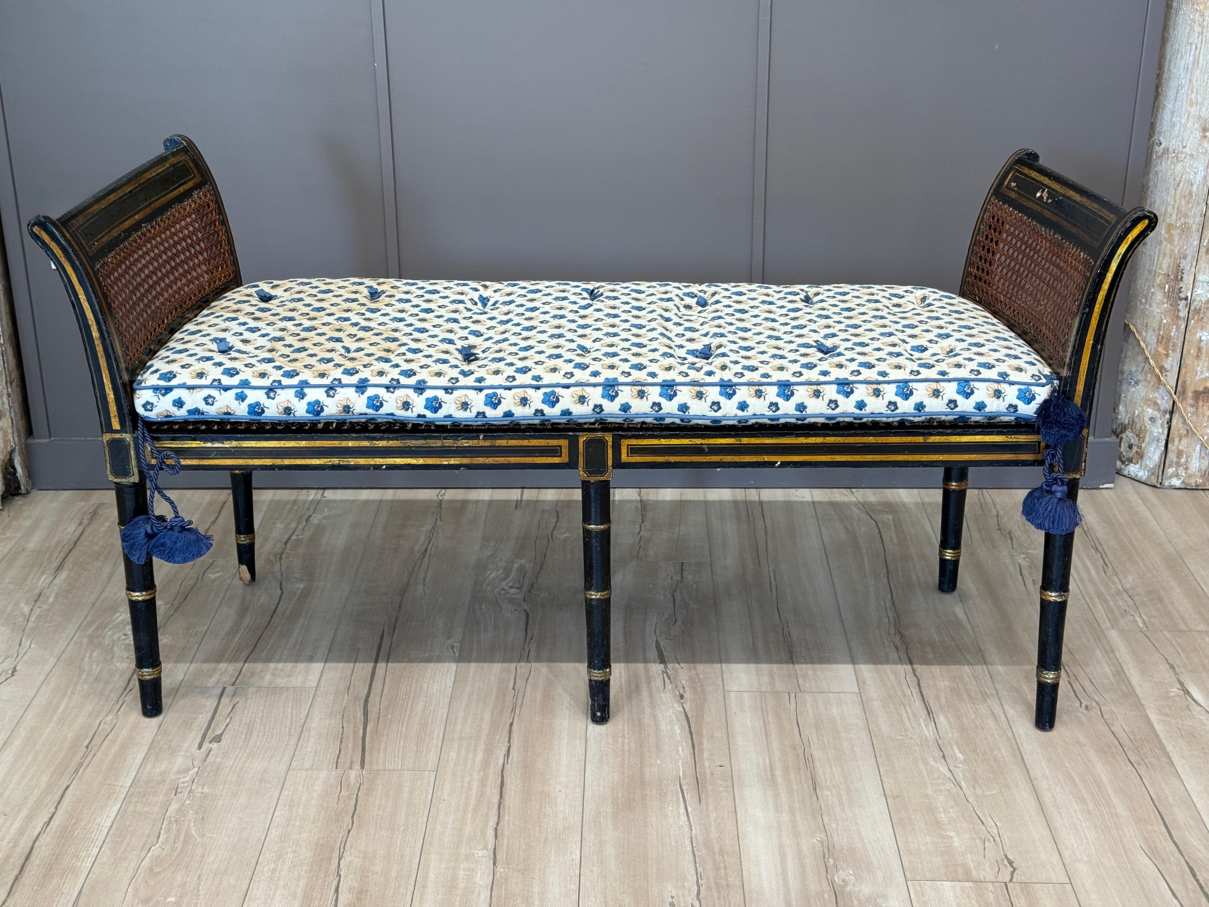 This English Regency period window bench features original black and gilt-painted surface with cane seat and ends, framed by curved supports and turned legs with gilt accents. The current cushion, done in a floral blue-and-cream print, is removable.