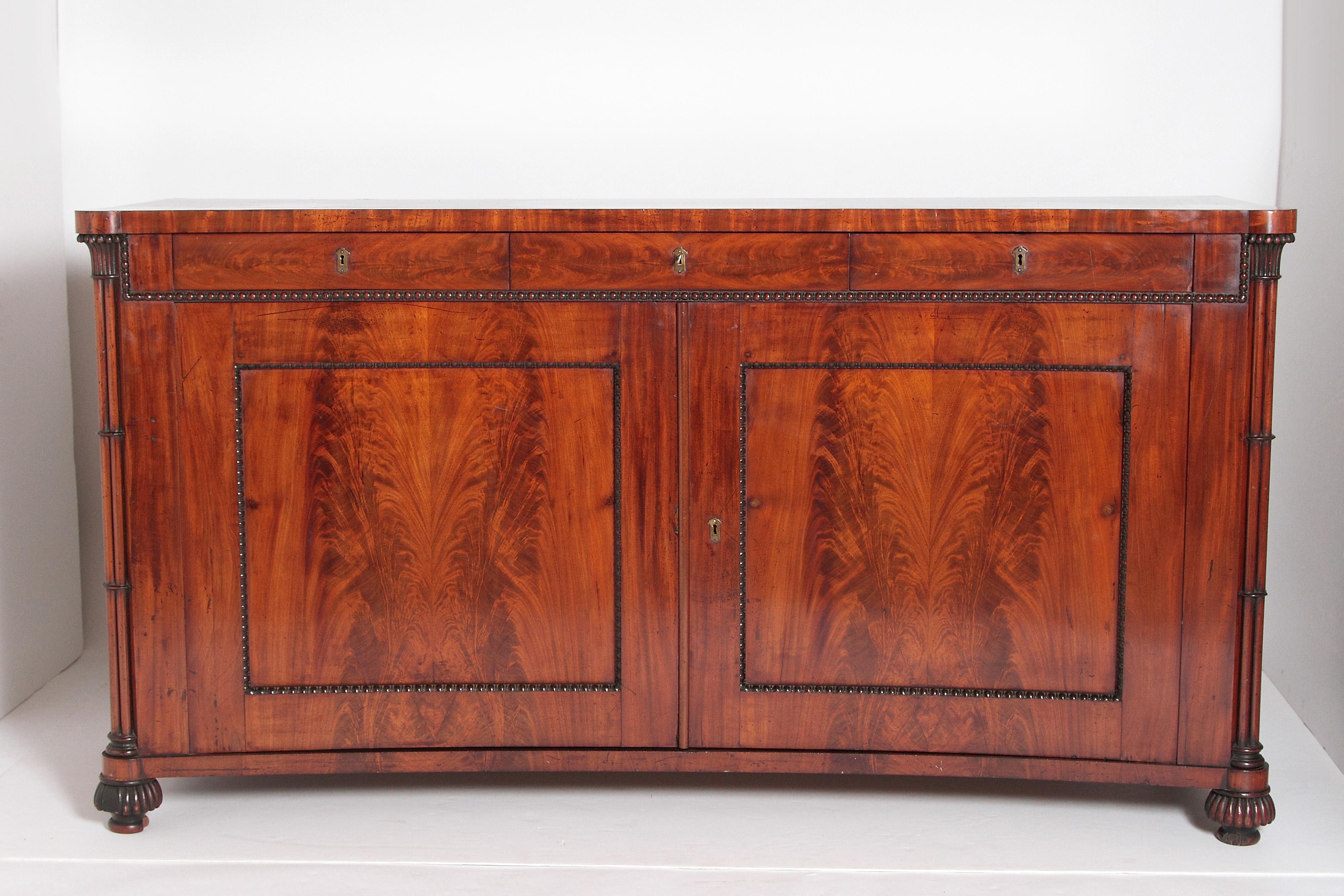 An English Regency mahogany cabinet with concave shaped top above three frieze drawers. Below are two large flame mahogany doors with carved bead borders. The interior contains four small drawers above one shelf. Reeded columns at the sides with