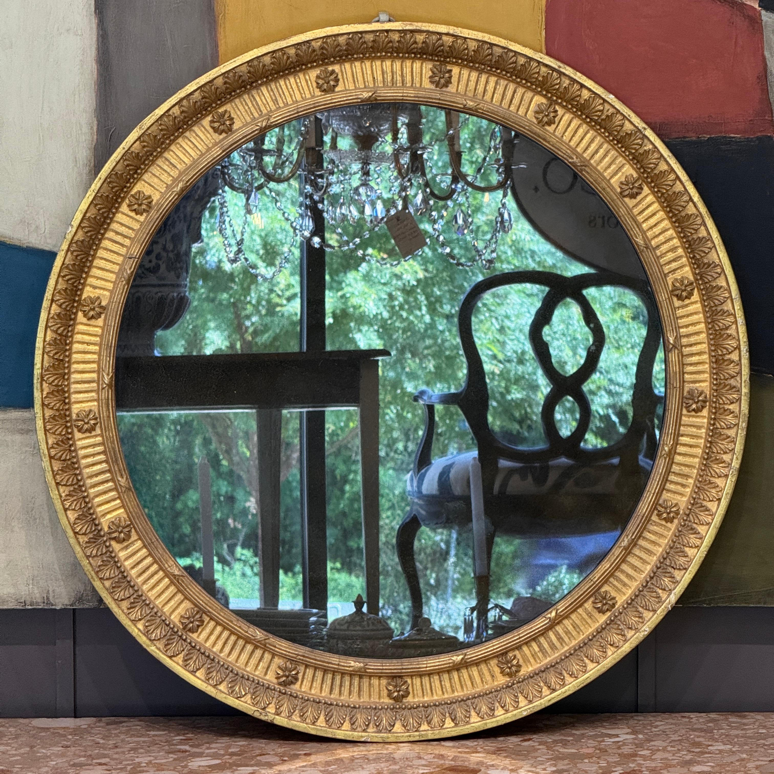 An early 19th century Regency period round mirror with carved giltwood frame featuring alternating acanthus leaf and floral medallion accents. The original convex mirror plate is set within a deeply molded and finely detailed surround, retaining its