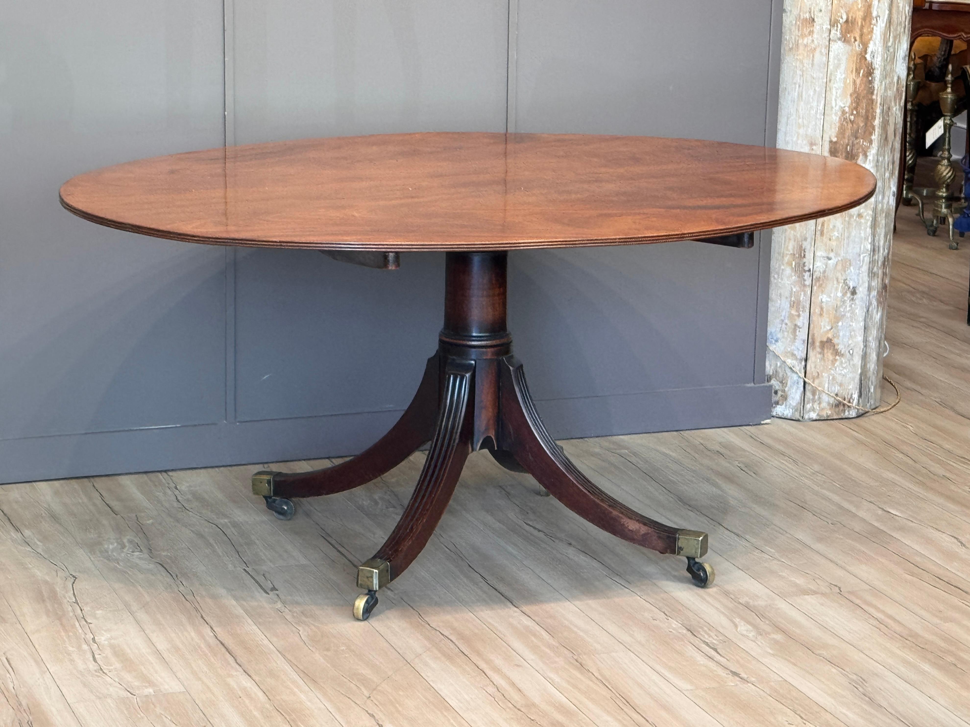 Table à déjeuner ovale à plateau basculant en acajou magnifiquement figuré, de style Régence anglaise du début du XIXe siècle. Le plateau est soutenu par un piédestal à balustre cannelé reposant sur quatre pieds évasés munis de chapeaux et de