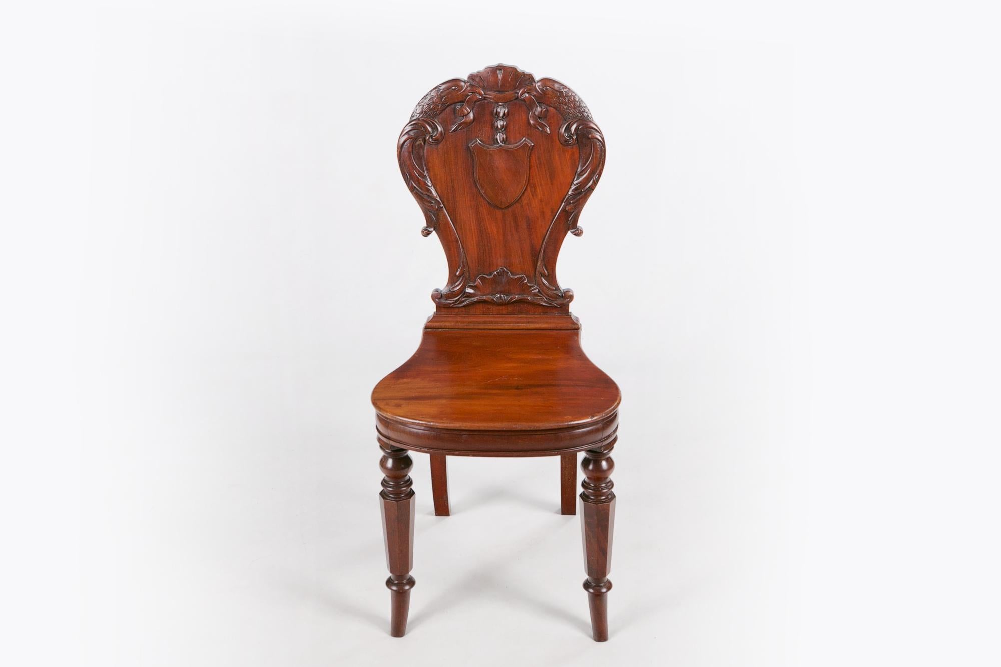 Early 19th century Regency pair of mahogany hall chairs by Gillows of London & Lancaster, the carved moulded back with centred armorial shield surmounted with foliate crest flanked with pair of phoenix birds with ribbon motif, all carved in high