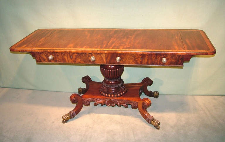 An unusual early 19th century Regency period figured mahogany sofa table having rosewood, maple, boxwood and ebony crossbanded top above angled frieze drawers raised on boldly carved and fluted centre pedestal with shell-carved platform base ending