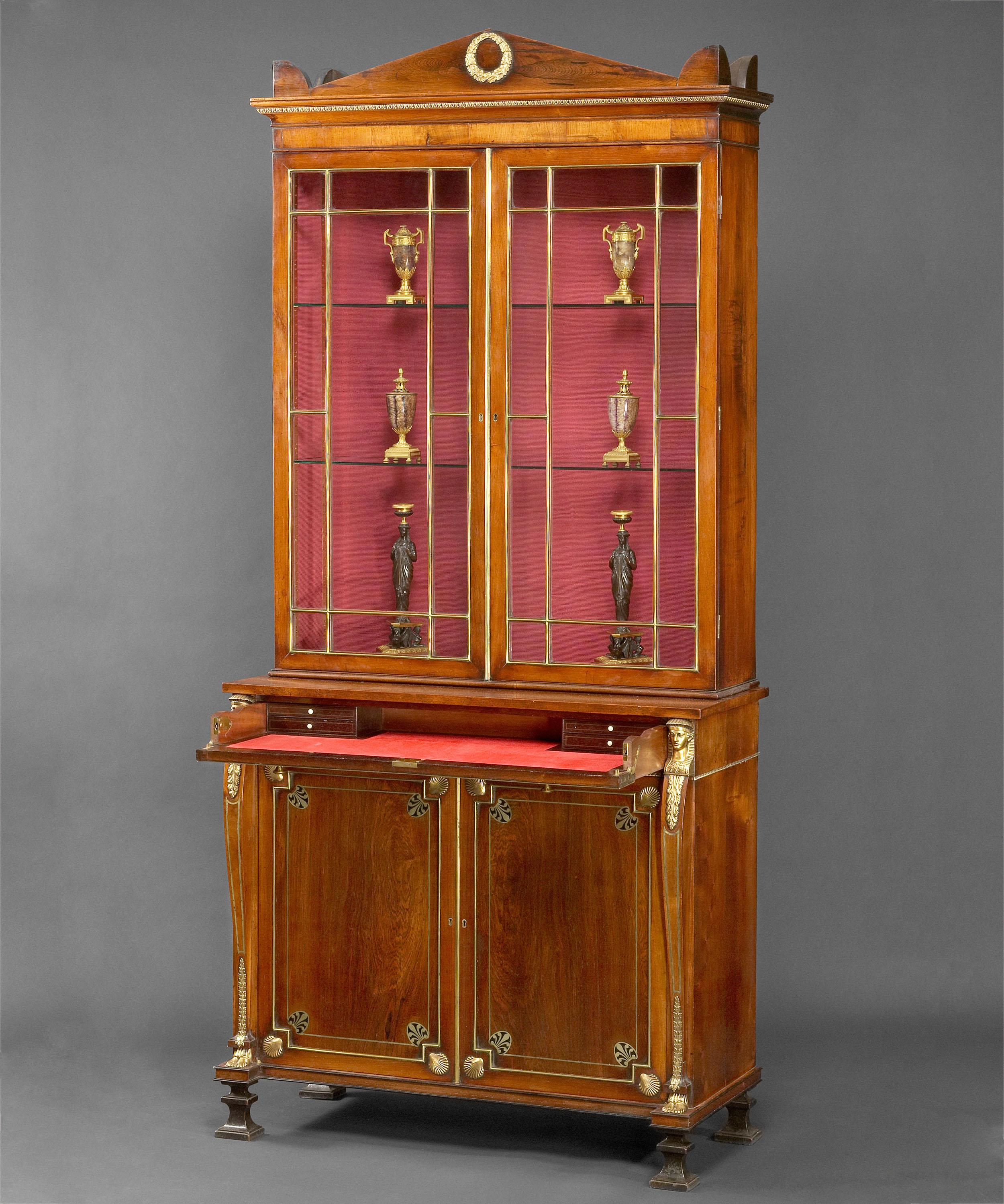 The acroteria and apex cornice, which has an ormolu mounted laurel wreath, rests on a moulded cavetto cornice with lappeted ormolu band above a pair of geometrically glazed doors with brass glazing bars. The base has a slim secretaire drawer mounted