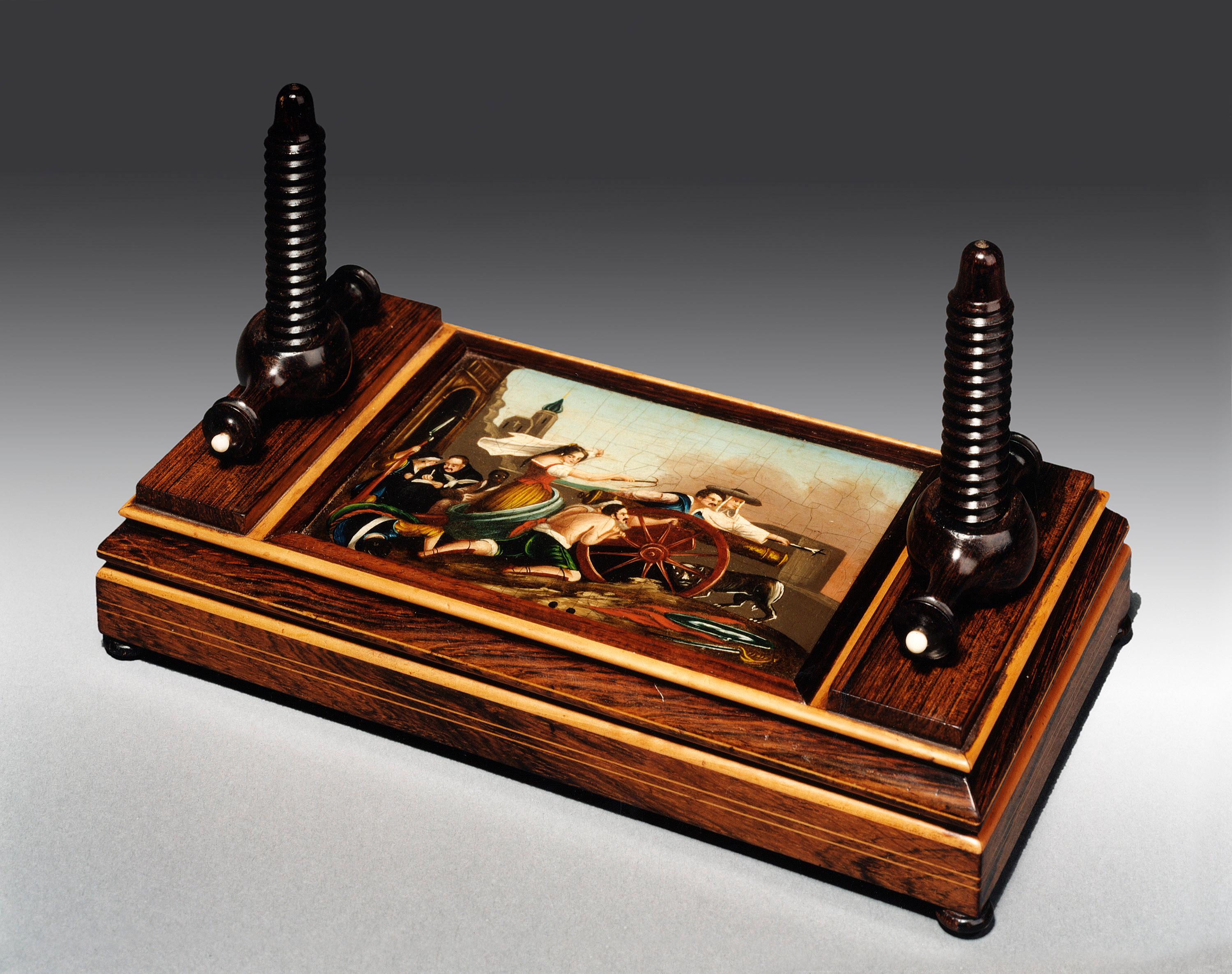 The two carved and turned wooden 'bolts' and 'nuts' secure the carved upper plate to the lower plate or base. The upper plate has a small painting entitled 'The Defence of Saragossa by Sir David Wilkie. The original painting was purchased by George
