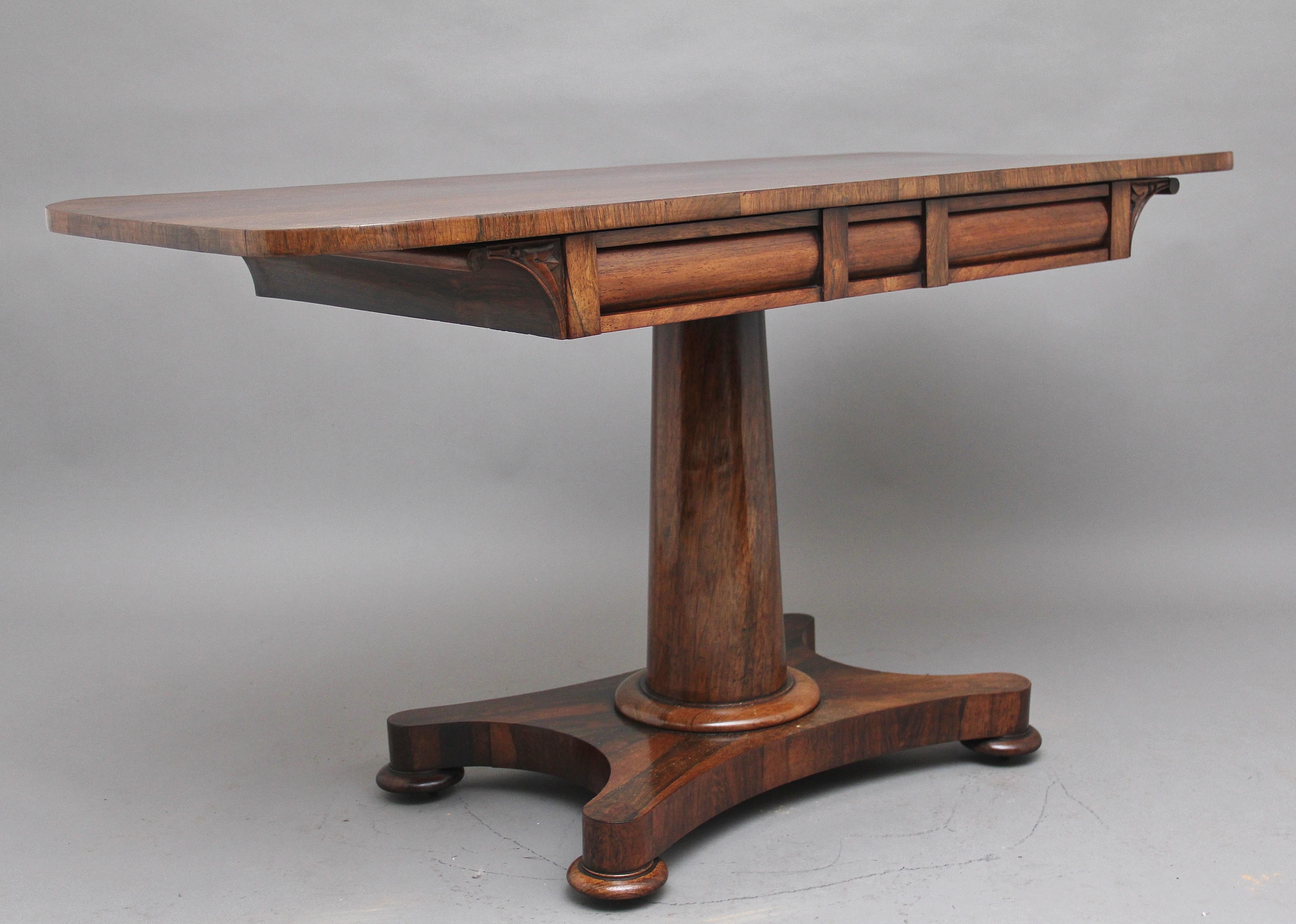 Early 19th century rosewood sofa / library table, having a lovely figured top of rectangular shape with rounded corners above two oak lined frieze drawers, supported on a turned column on a shaped quatrefoil platform base raised on bun feet, circa