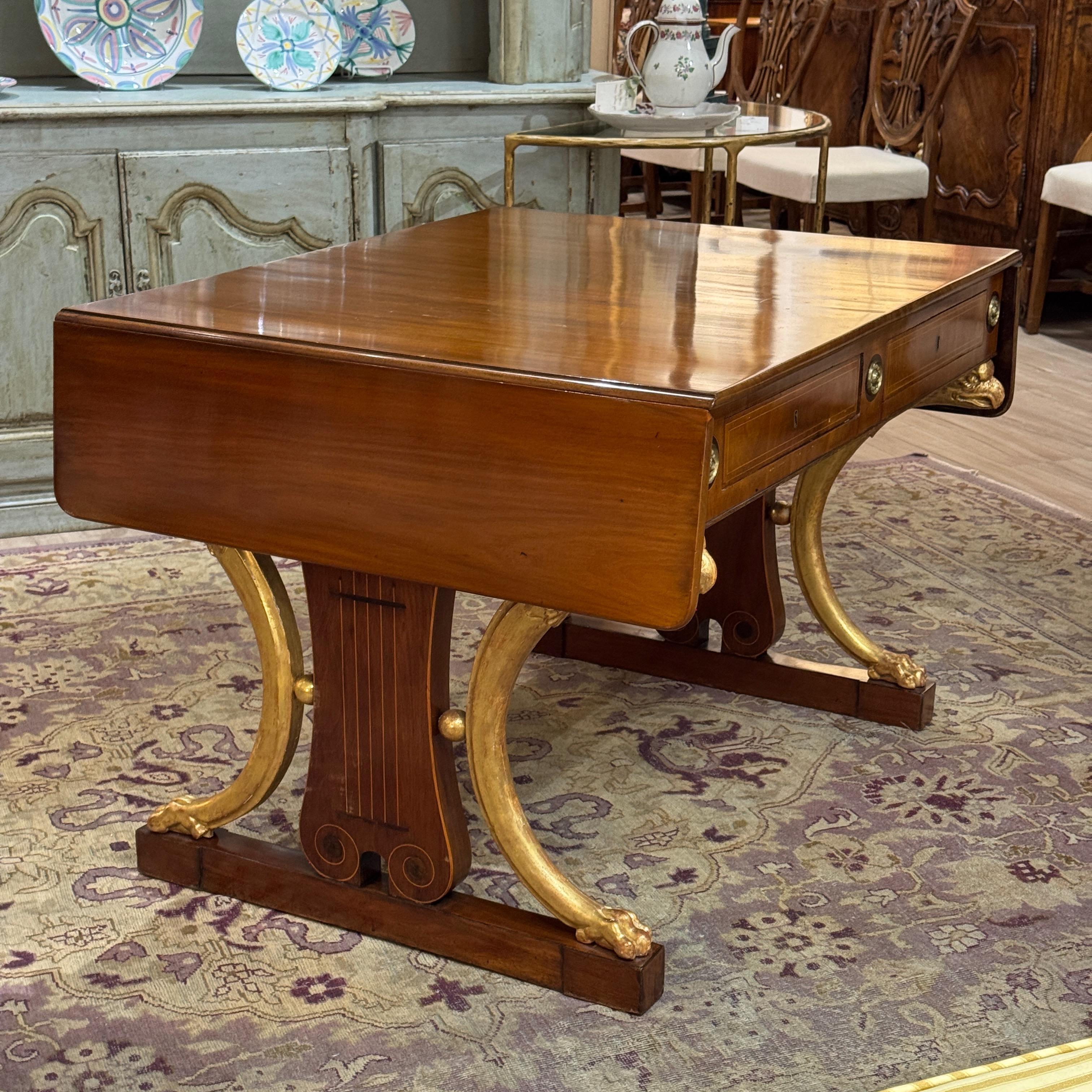 Rich mahogany and bold parcel-gilt supports define this early 19th century Russian sofa table. The lyre-form ends feature gilt scroll legs terminating in paw feet, while the apron houses inset drawers with brass roundels and fine line inlay. Elegant
