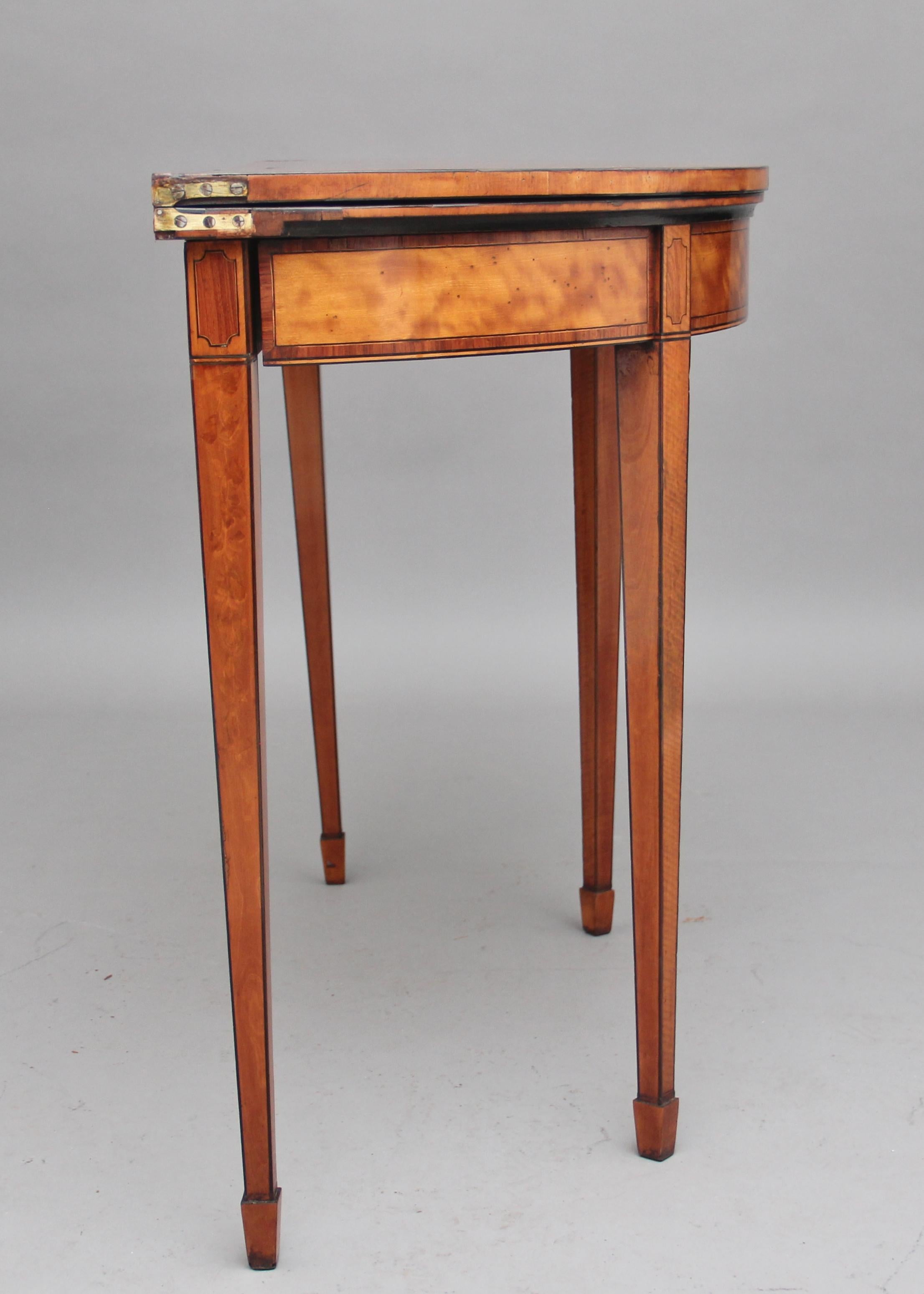 Early 19th century satinwood Demilune card table, the semi-circular crossbanded top opening to reveal a green baize playing surface, the frieze below also crossbanded and having an inlaid decorative pattern at the top of the legs, supported on four