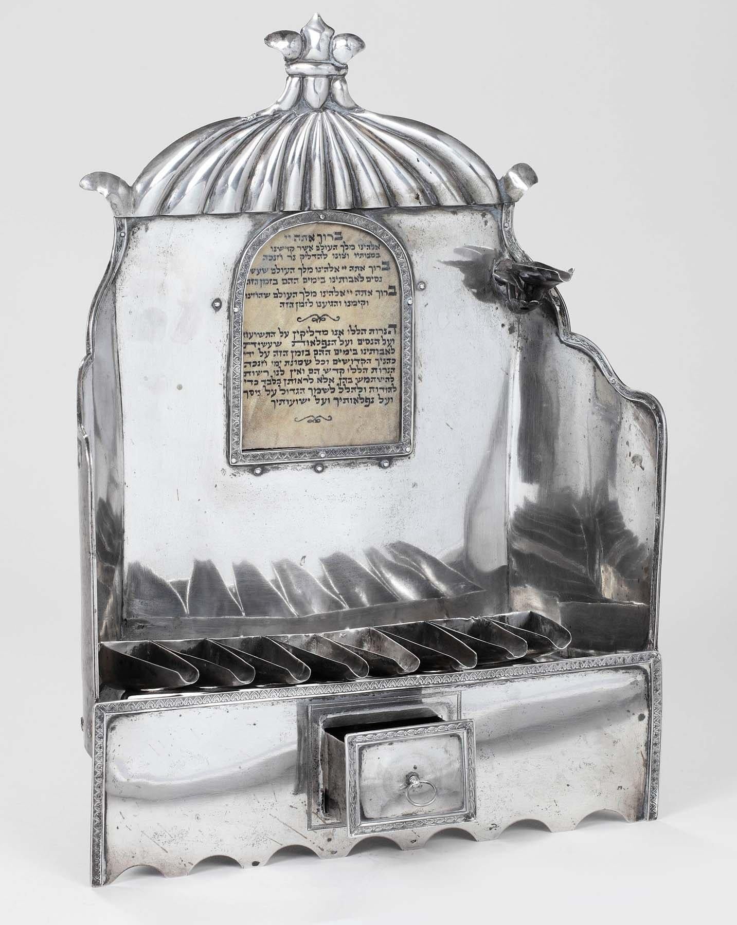 This large silver Hanukkah lamp features a canopy at top, with a cut-out window below where a piece of parchment could be inserted that would have the blessings of Hanukkah inscribed on it (the parchment is a later replacement). Row of oil fonts