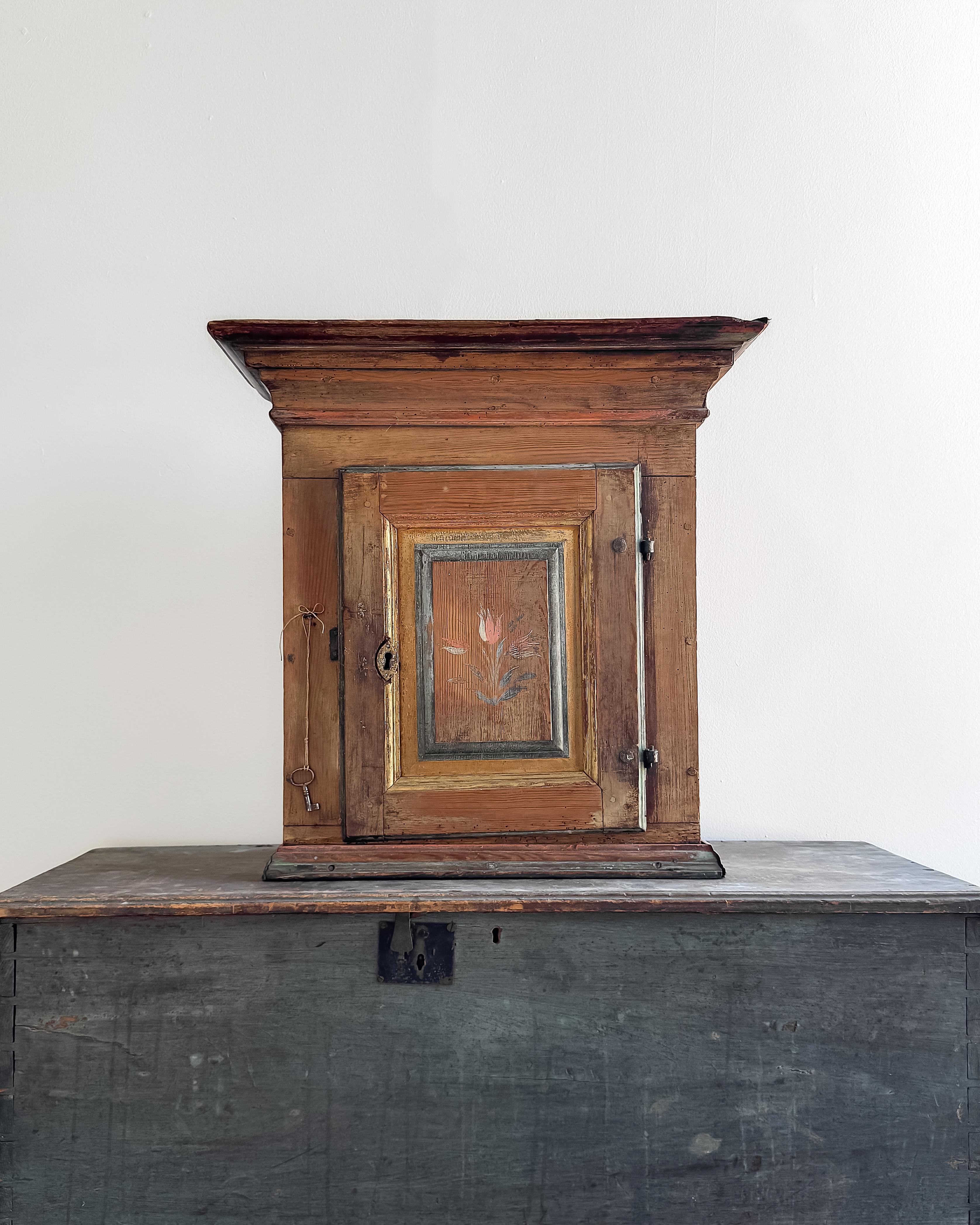 Chargée d'histoire, cette petite armoire murale suédoise présente une magnifique tulipe peinte à la main sur la porte, encadrée par des couches de peinture vieillie dans des tons doux et sourds. 

L'extérieur a conservé sa finition usée d'origine,