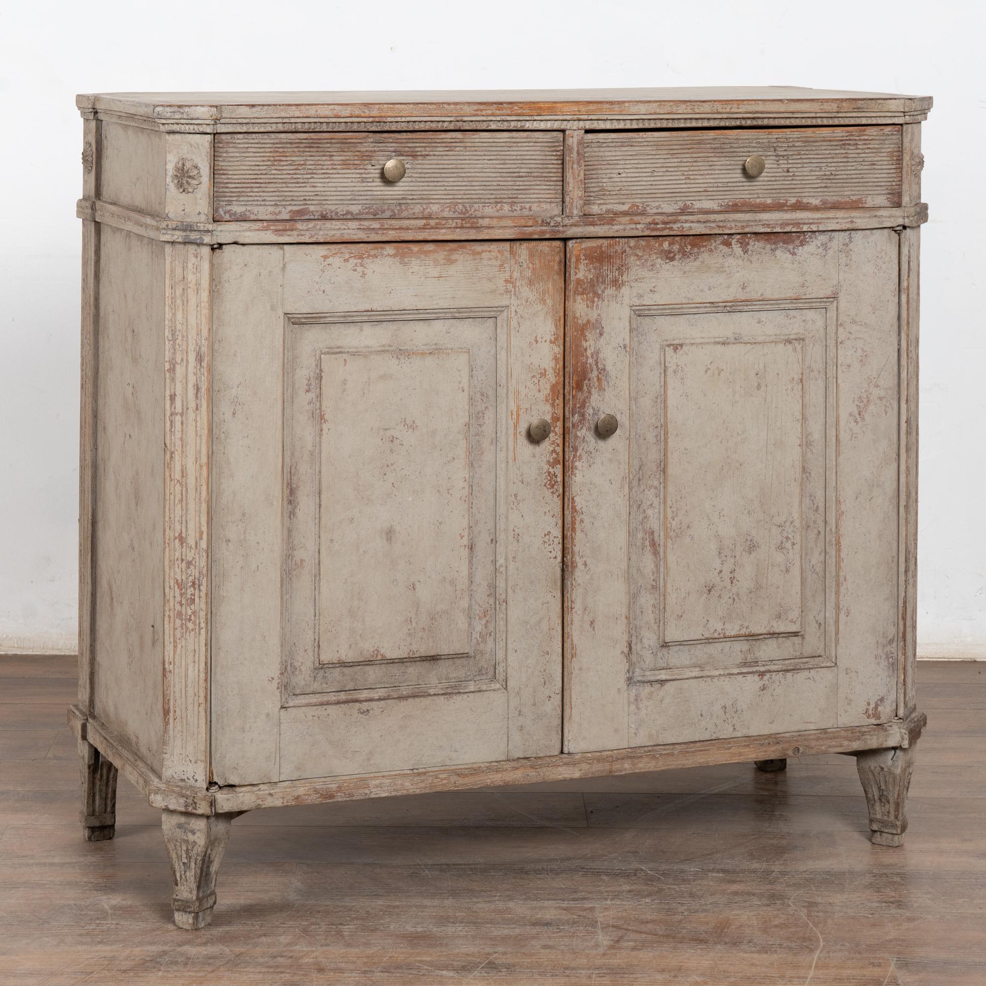Questa credenza di campagna svedese ha una finitura verniciata in grigio invecchiato che completa il tradizionale intaglio scanalato lungo i cassetti e i lati inclinati, che poggiano su piedi affusolati.
I due cassetti e le ante si aprono con