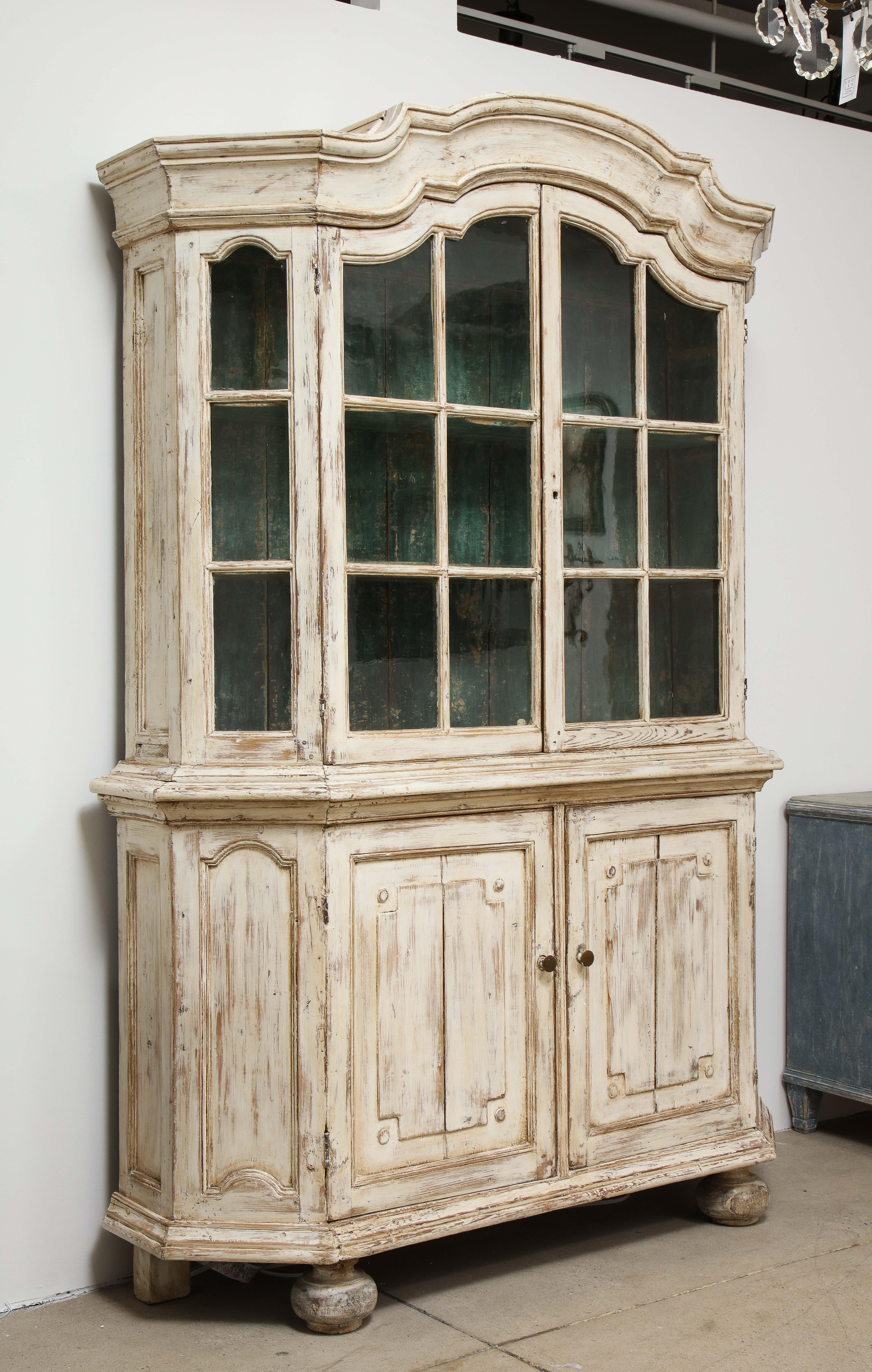 An antique Swedish vitrine in the Gustavian style painted in an ivory color. The top features glazed doors with glazed side panels and a green painted interior. The base has two doors that open to reveal a single shelf. The base rests on bun feet.