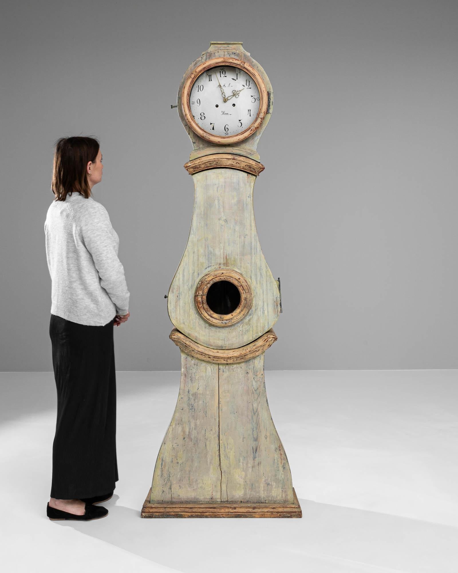 This early 19th Century Swedish wooden floor clock stands as a towering testament to the enduring elegance of Scandinavian design. Its tall, slender body is shaped from wood that bears the gentle patina of time, its surface graced with the faded