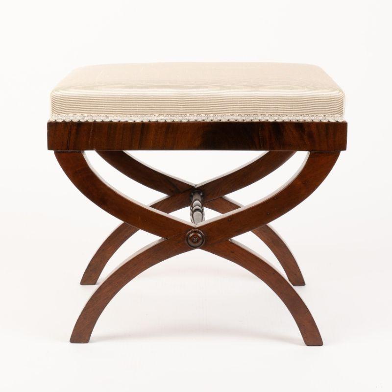 Mahogany curule stool with arched crossed legs joined at the pivot point by turned stretcher. The stretcher is defined by turned rosettes at the insertion into the leg crossing point. The legs support an upholstered boxed seat on a mahogany cross