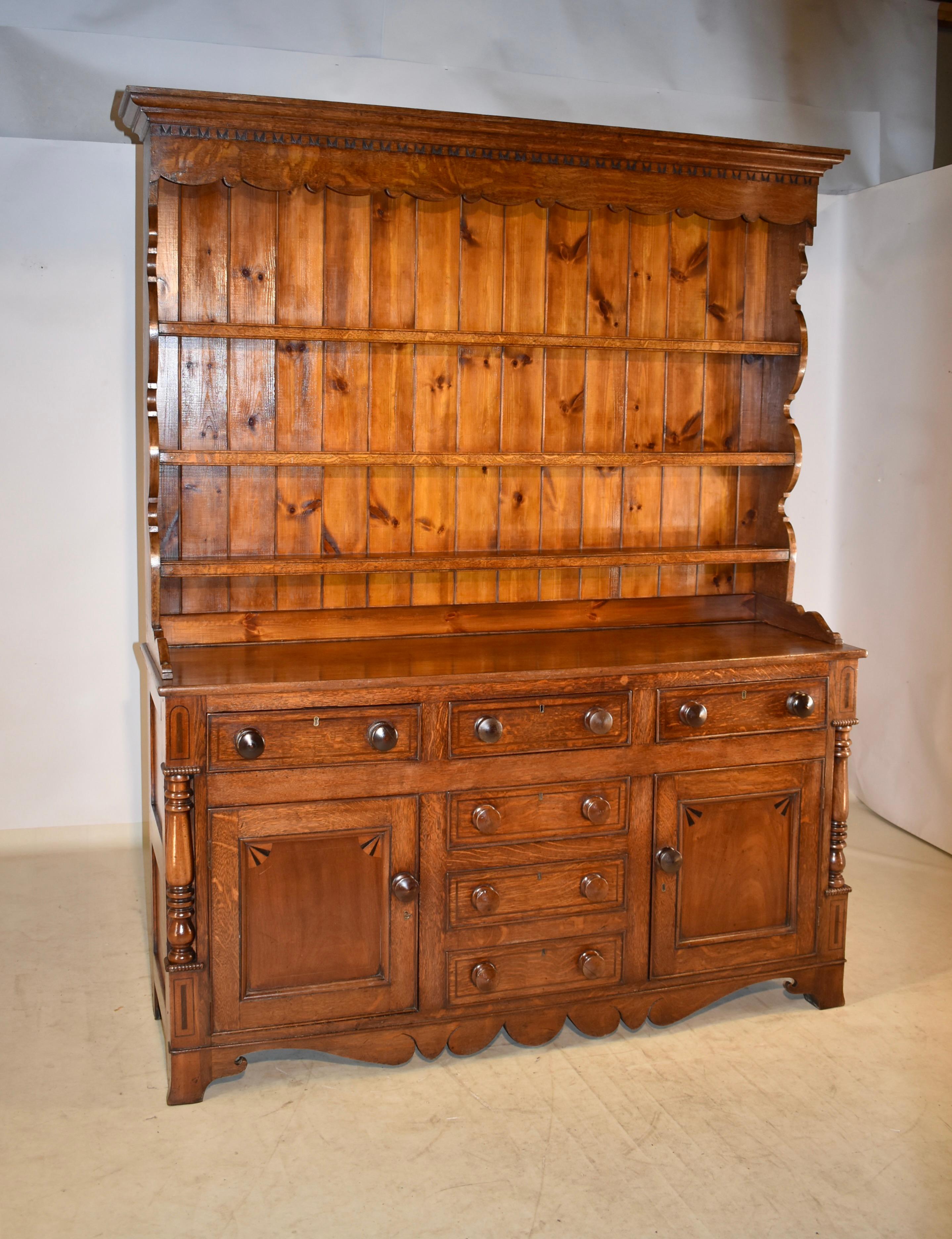 Early 19th century welsh oak dresser with crown molding gracing the top of the cabinet over a scalloped and hand carved decorated apron. It has three shelves which are flanked by scalloped sides. The top sits atop the base, which has paneled sides