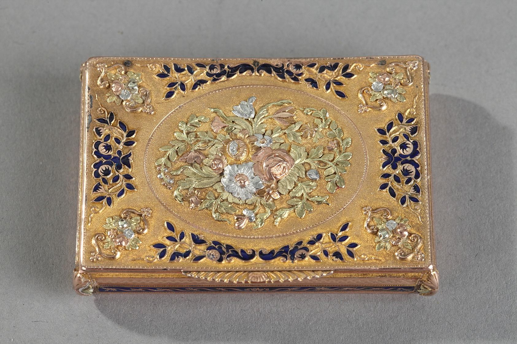 Rectangular box with three tones of gold and royal blue enamel. The hinged lid is embellished with a multicolored gold medallion in relief featuring a bouquet of roses and tulips, set on a matte gold background. Delicate foliage accented with royal