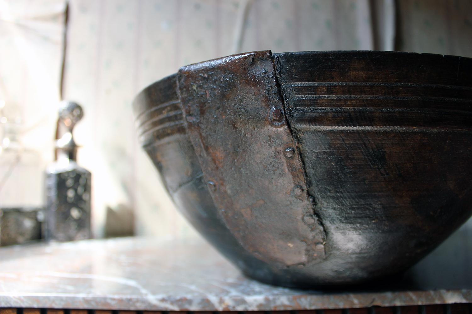 The wonderful treen bowl, in beech, having a residual dark stain, groove turned rim band and a lovingly executed blacksmith sheet metal patch repair to one flank to mend a crack, the whole with a lovely deep patination and surviving from the second