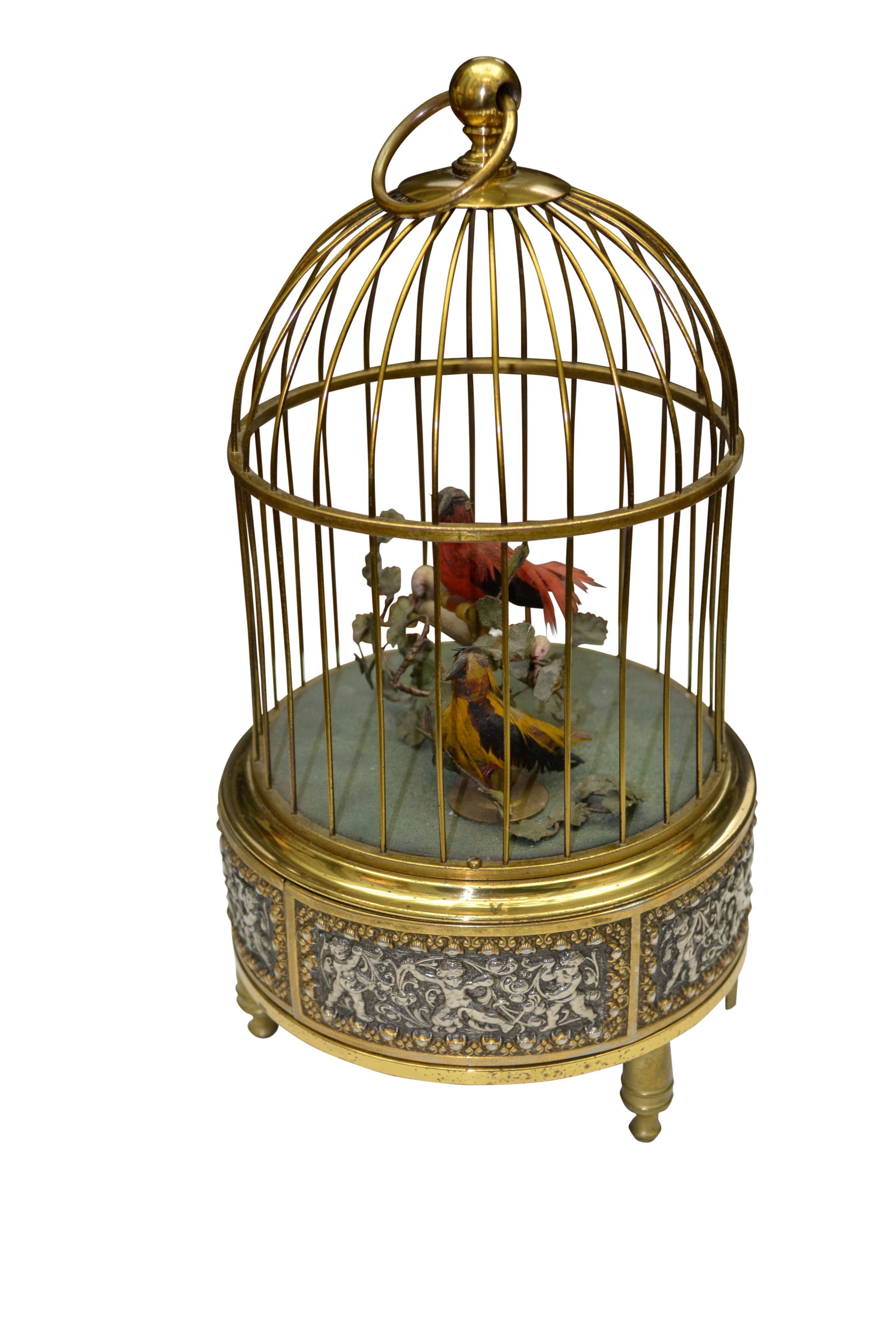 A brass wire cage with knob finial and loop handle; set on a slivered brass base with an ornate repousse frieze of frolicking putti; the whole supported by three turned brass feet; the interior has 2 animated manually feathered singing birds, one