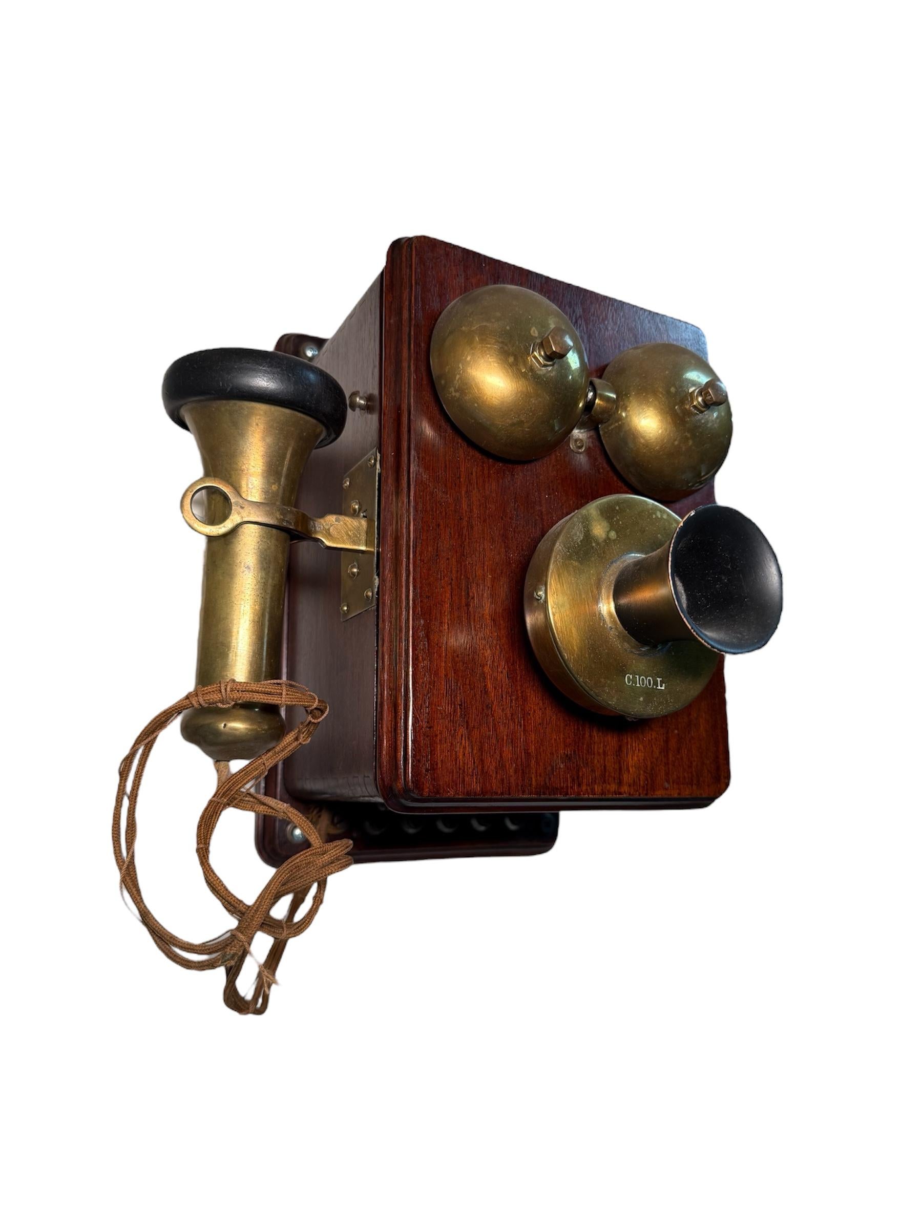 Si tratta di un telefono da parete dei primi del Novecento. Raffigura una scatola rettangolare in legno marrone scuro con campanelli in ottone dorato nella parte anteriore, cornetta su un lato e manovella sull'altro.