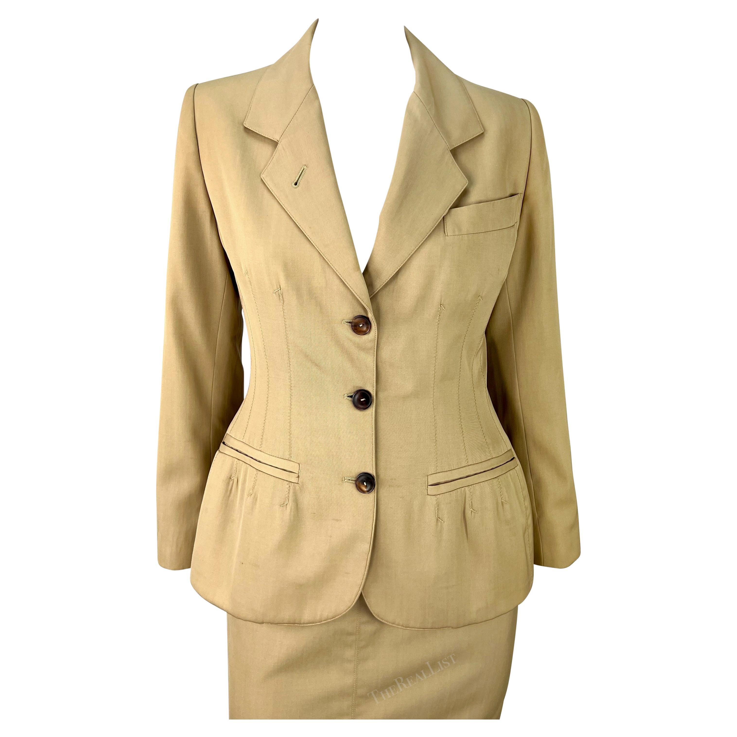Presenting a sleek beige Jean Paul Gaultier skirt suit. From the early 1990s, this suit consists of a fitted blazer and matching pencil skirt. The blazer features a peak lapel, darts at the waist, and light padding at the hem.

Approximate
