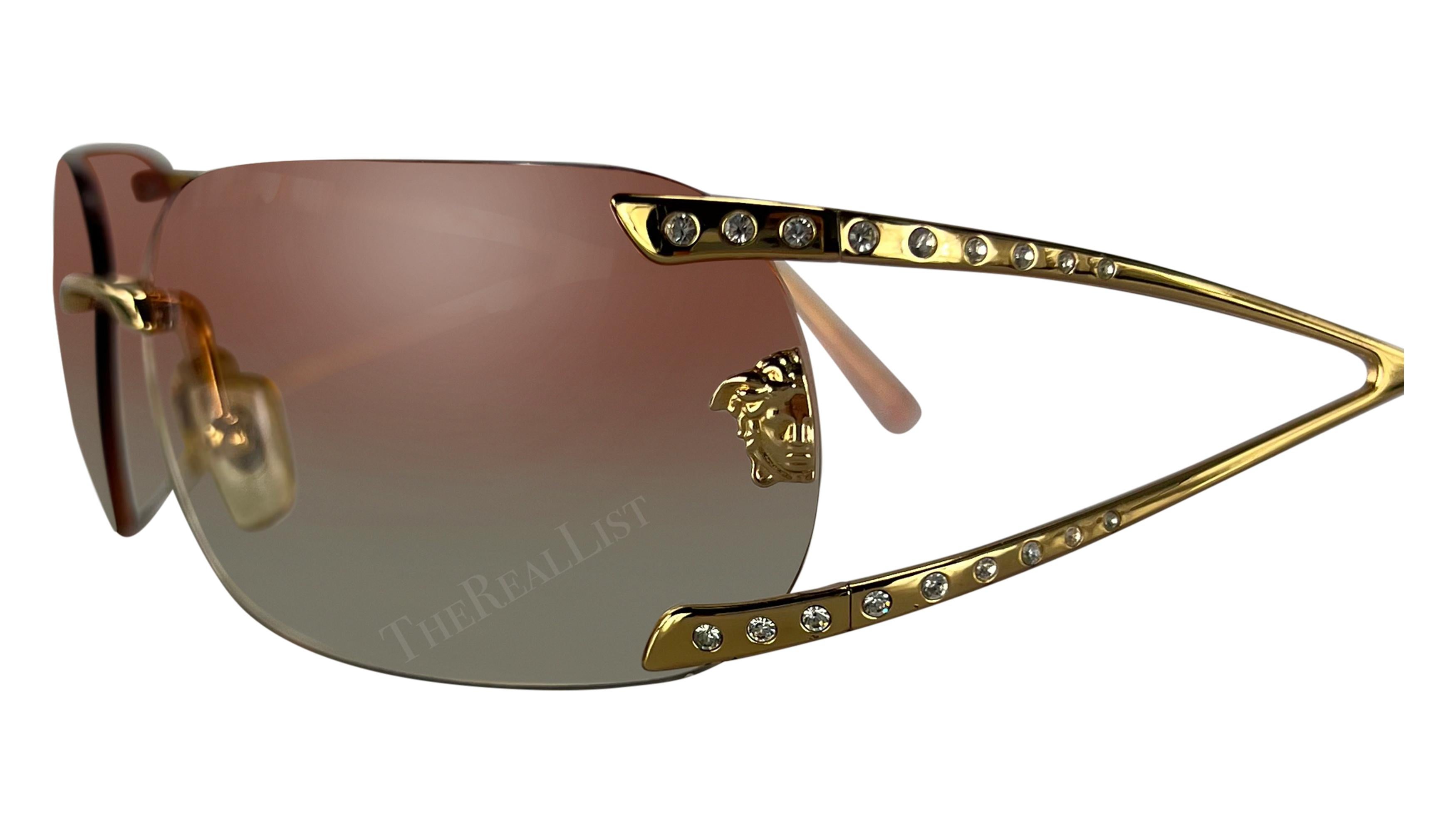 Datant du début des années 2000, ces lunettes de soleil oversize sans monture Versace, conçues par Donatella Versace, sont dotées de verres roses ombrés et de branches dorées rehaussées de strass. Elles sont complétées par un logo Versace Medusa sur