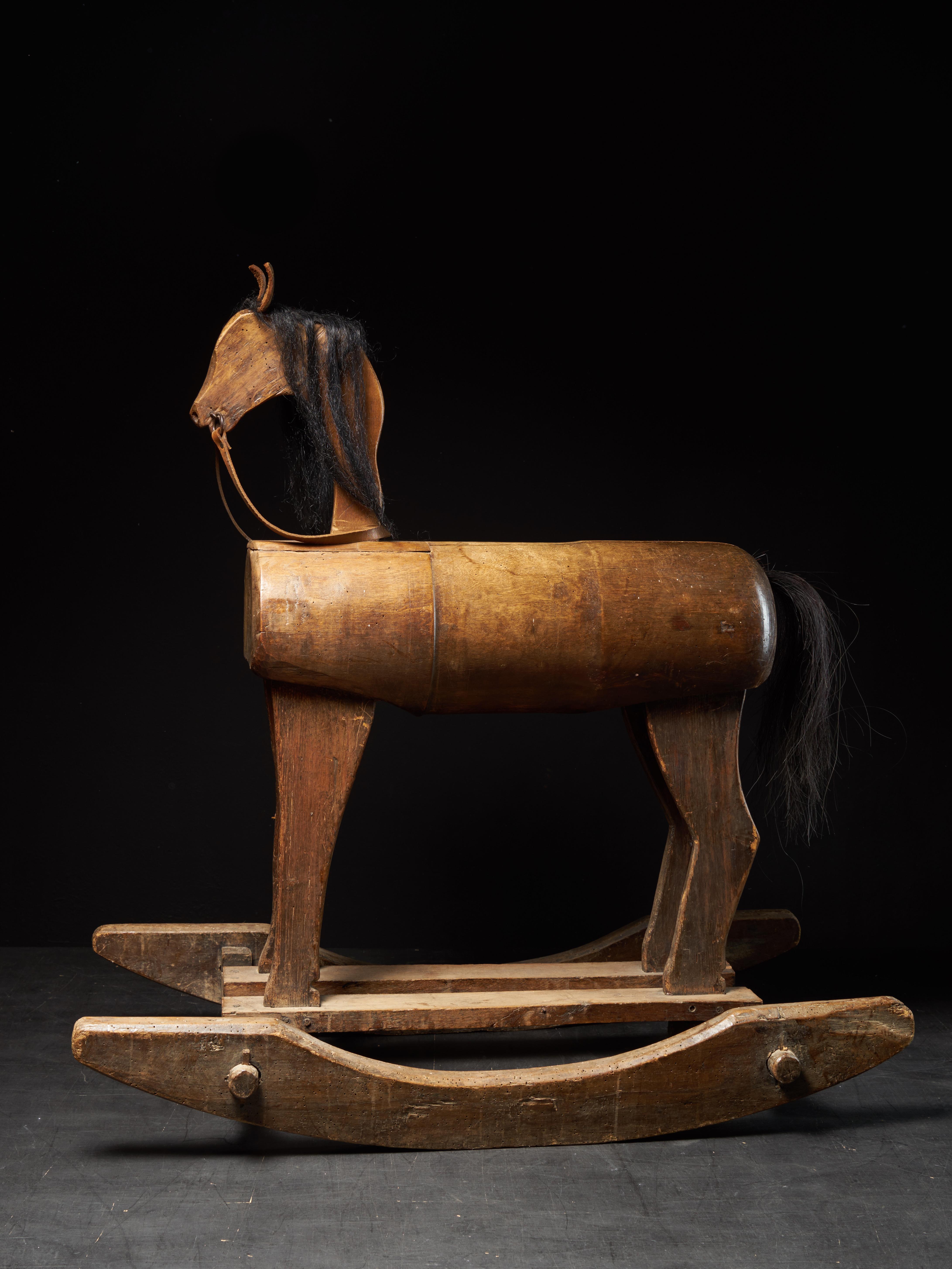 Le cheval à bascule a été fabriqué dans la première moitié du 20e siècle au Royaume-Uni. Les oreilles et les rênes sont en cuir brun clair. La tête et la queue du cheval sont faites de vrais poils et le corps, les jambes et la partie inférieure sont