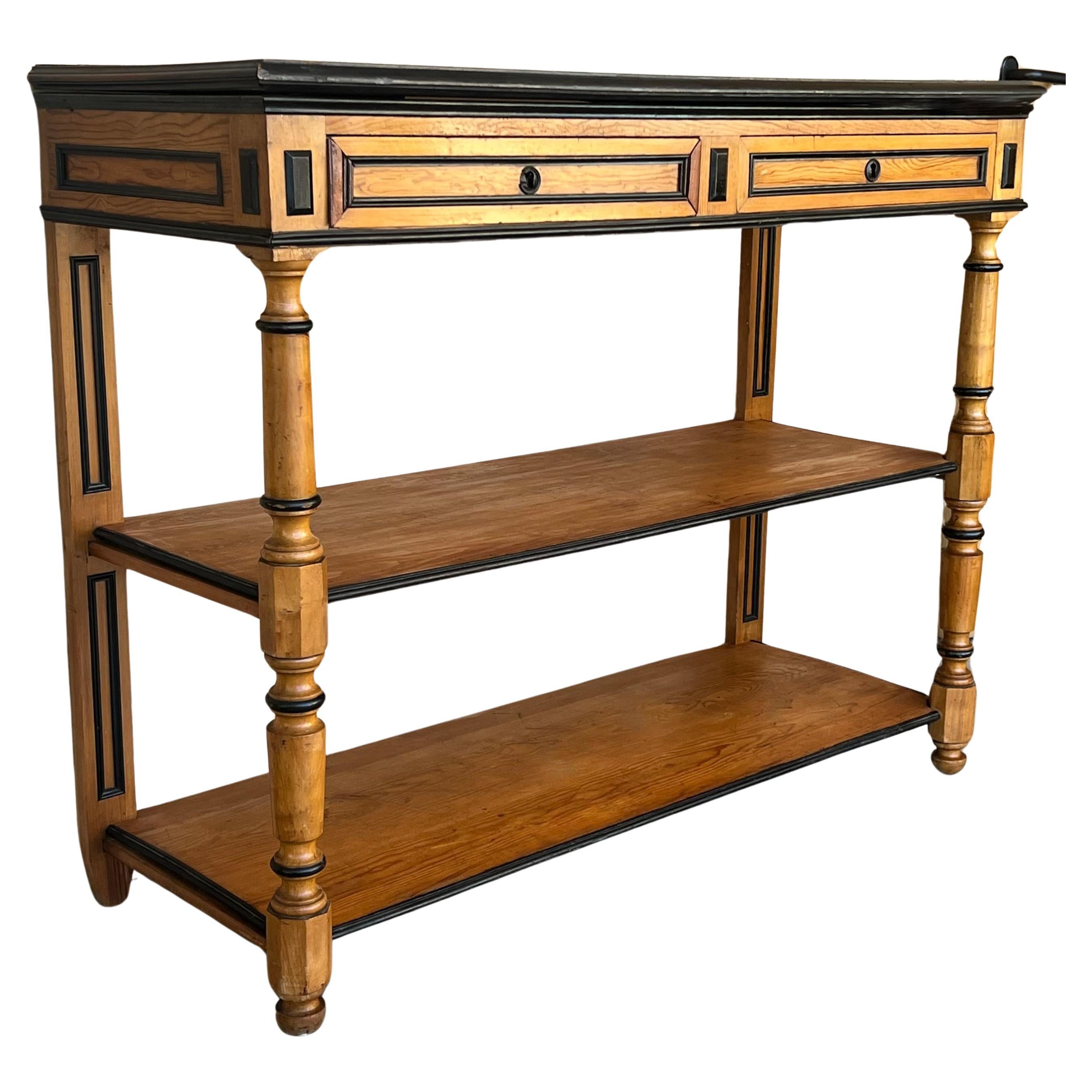 Trouvée en Angleterre, cette table de service en bois fruitier datant de 1900 est dotée d'un plateau avec deux tiroirs munis d'une serrure fonctionnelle et de deux étagères à l'étage inférieur. Pieds tournés décoratifs à l'avant, joliment ébonisés