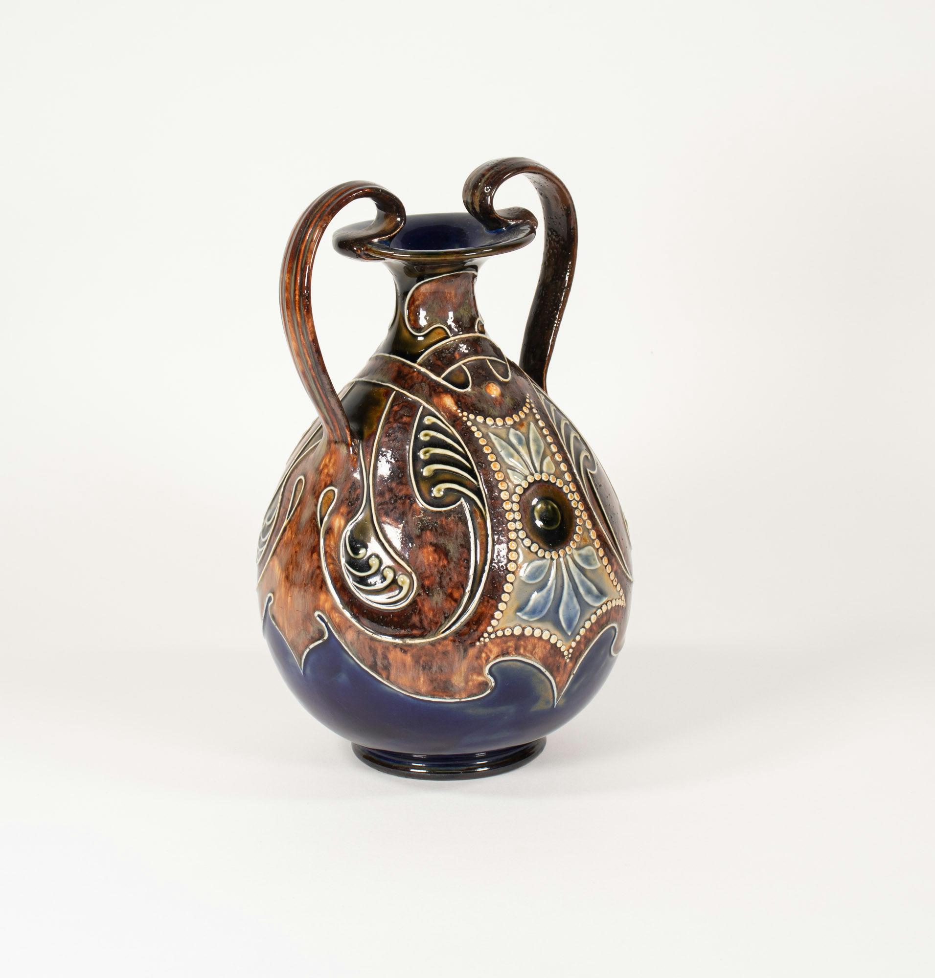 Quest'anfora tardo-vittoriana in gres inglese Arts & Crafts Royal Doulton Lambeth di Frank A. Butler è stata realizzata con il modello n. 960. Questo vaso a due manici è decorato con uno smalto screziato color ambra bruciata e altri elementi più