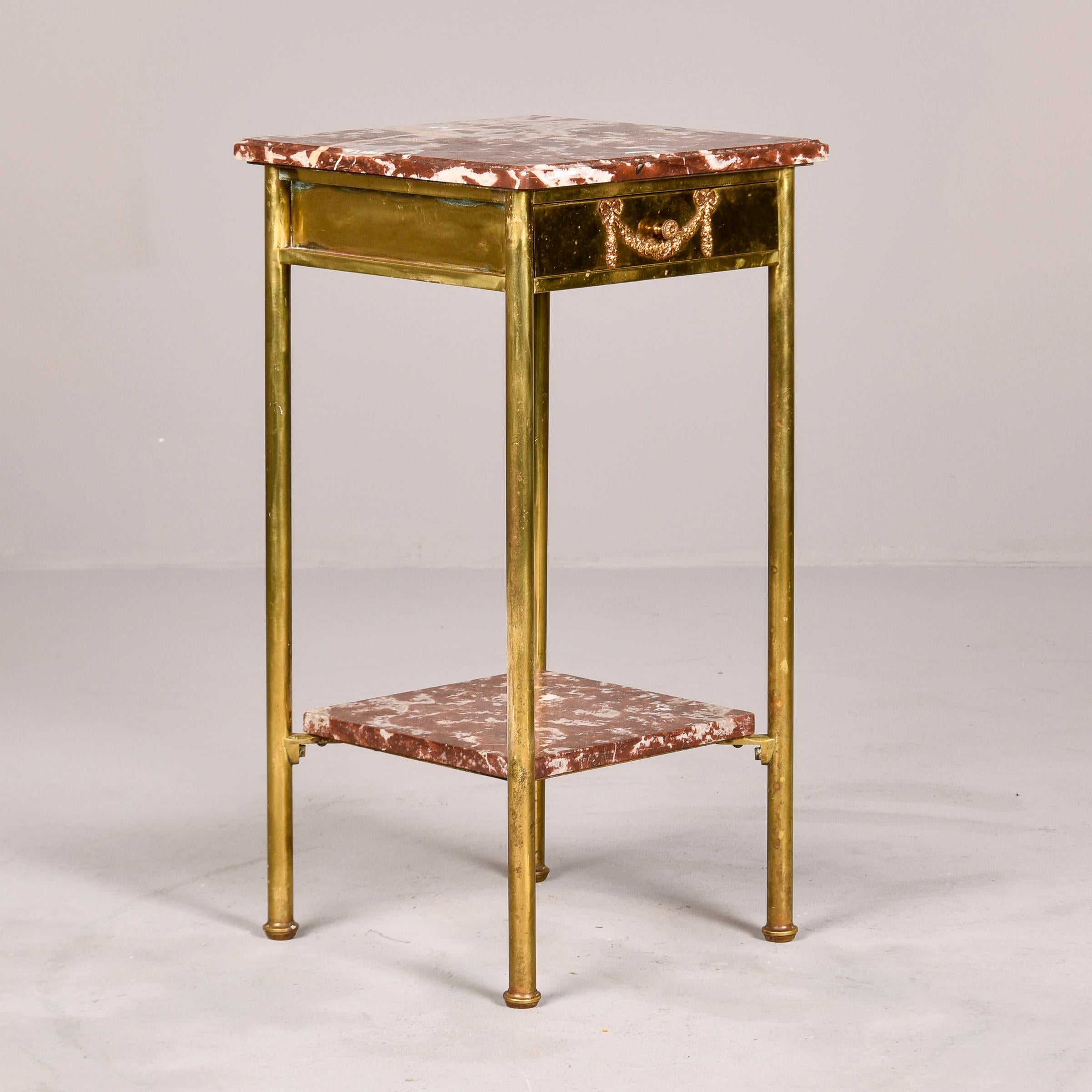 Circa 1910 French brass side table with marble top and marble lower tier. Small center drawer has dovetail construction and decorative repousse detail with bow-topped swag. Brass legs and marble top and lower shelf in shades of rusty brown with