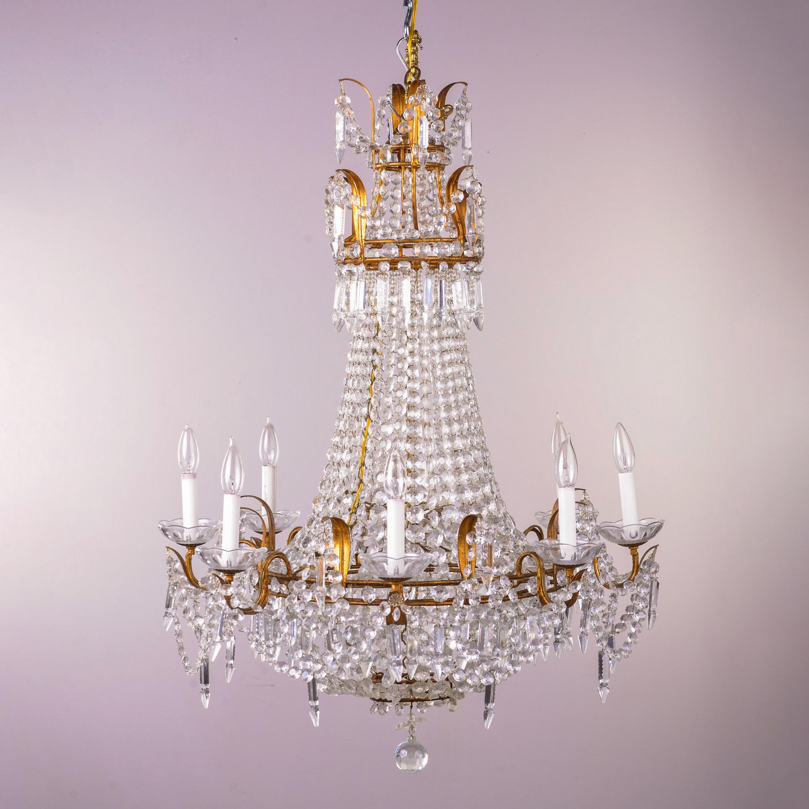 Found in France, this circa 1920s empire style chandelier features a basket form lower tier ringed with eight candle style lights with candelabra sized sockets. The top tiers and base have gilt metal leaf form accents and are draped in strings of