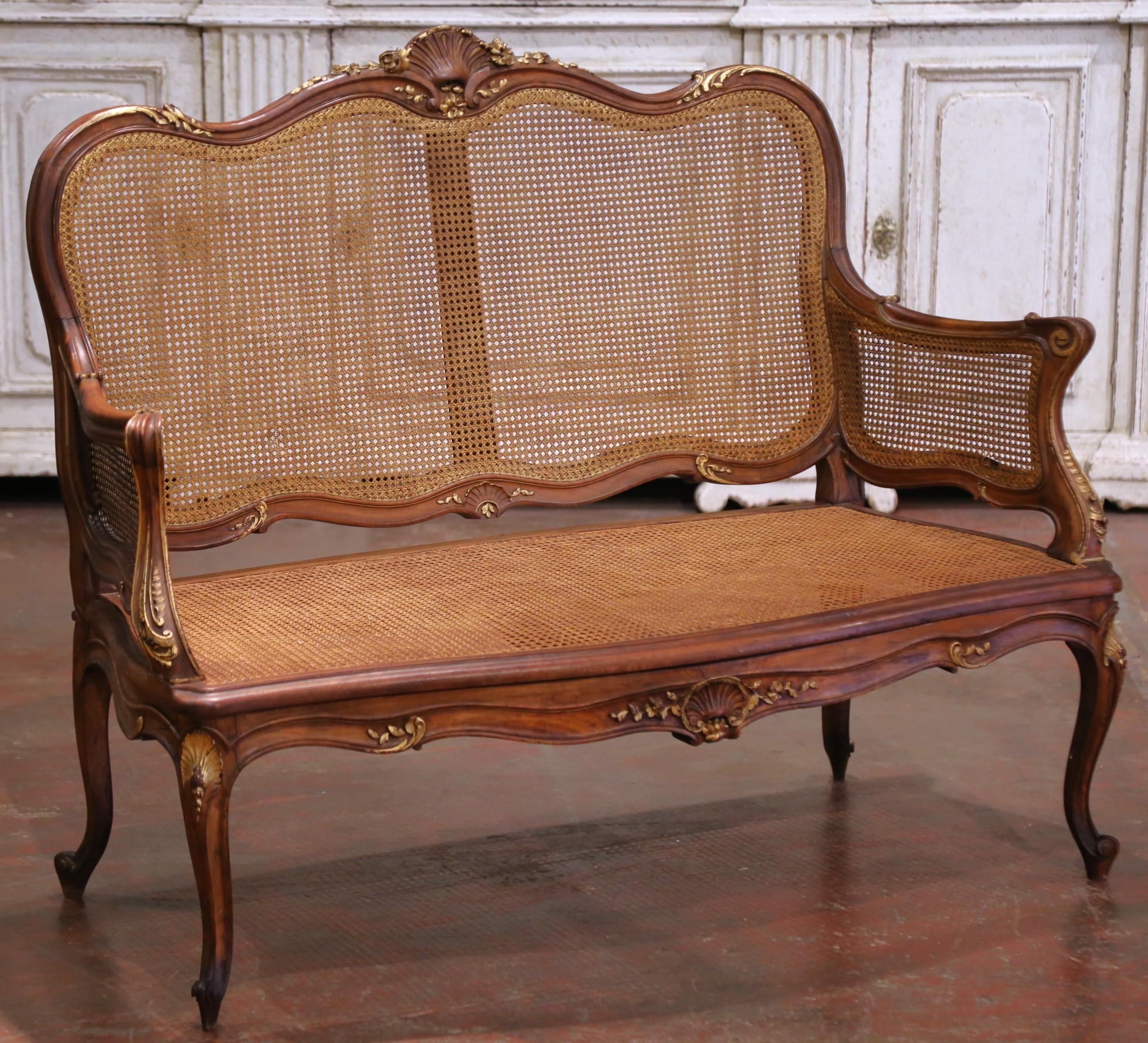Elegant and finely detailed, this French Louis XV style settee was crafted circa 1920 from richly patinated walnut with subtle parcel gilt accents. The shaped frame features a carved shell crest and scalloped serpentine rail over a tight cane back,