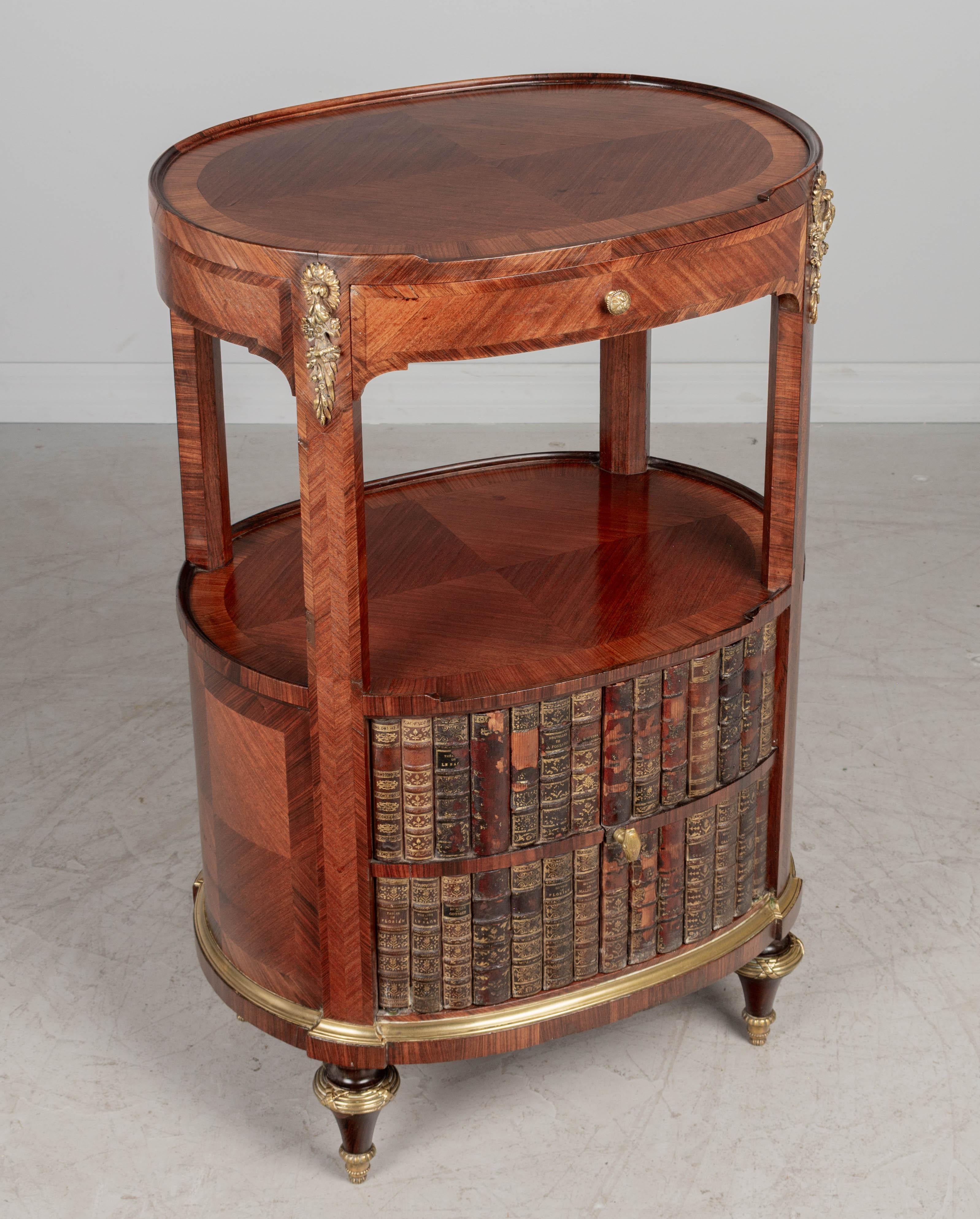 A fine early 20th century French Louis XVI style oval marquetry side table with faux livres doors. Made of mahogany with rosewood veneer and finished on all sides. Good quality cast bronze decorative hardware. Gold embossed leather book spines are