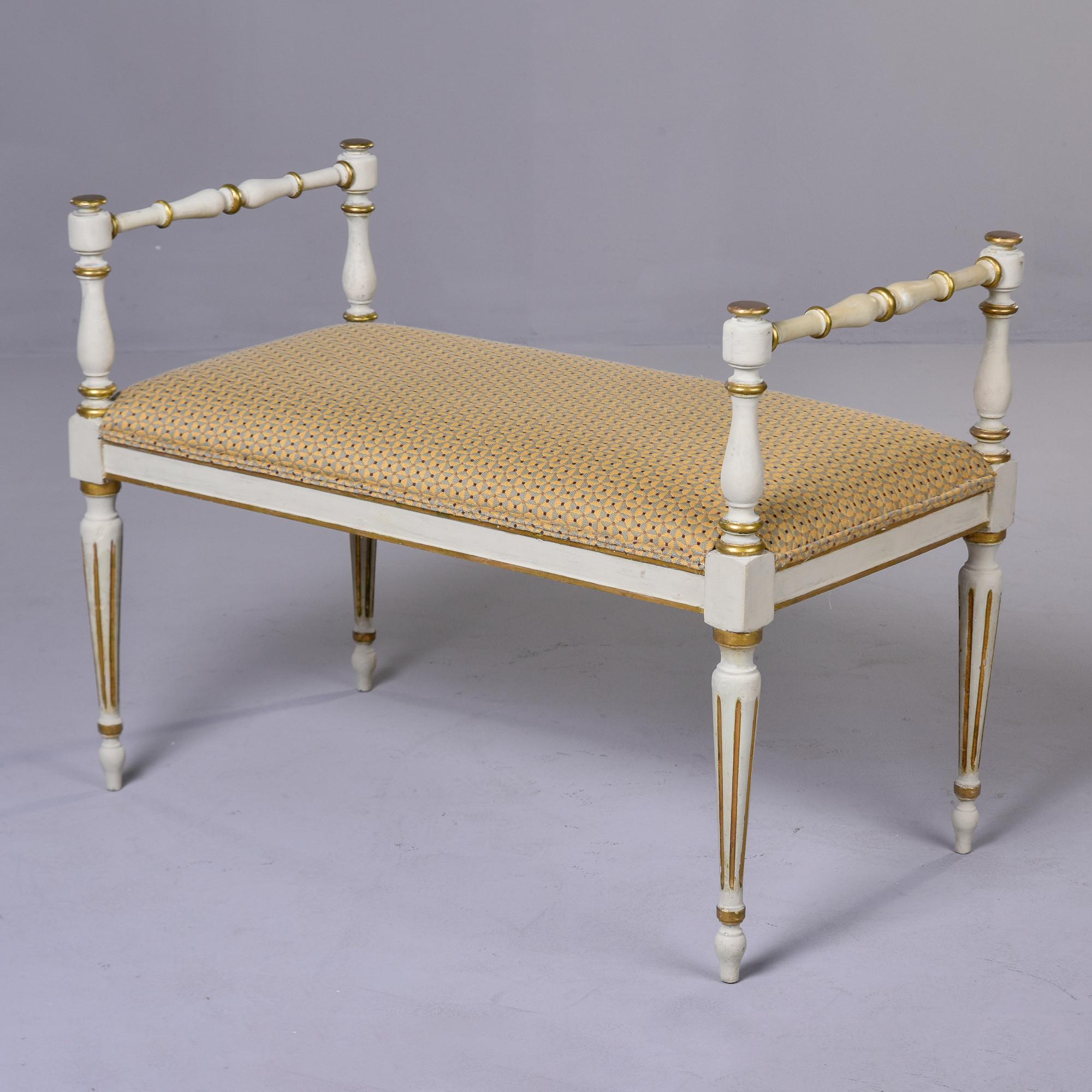 Found in France, this circa 1920s upholstered bench has an antique white painted frame with gilded detailing. Frame has arms with turned details, gilded accents and traditional tapered and reeded French legs. New upholstery in gold and blue gray