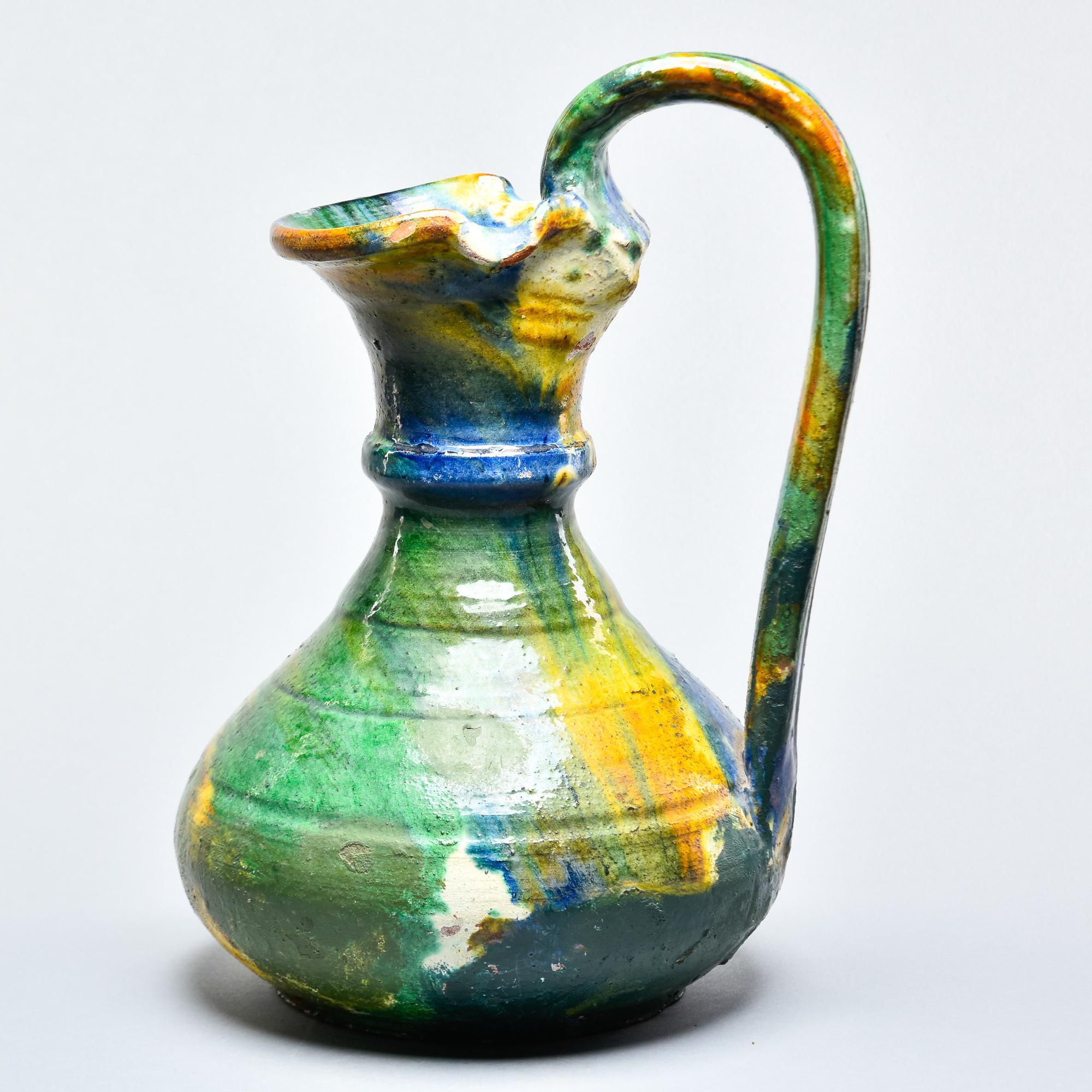 Found in France, this hand thrown ceramic French ewer or pitcher dates from approximately 1910. This piece stands 14.5” high and has a tall, curved handle and flared spout with a wide vessel body. Green, blue, gold and cream colored glaze on the
