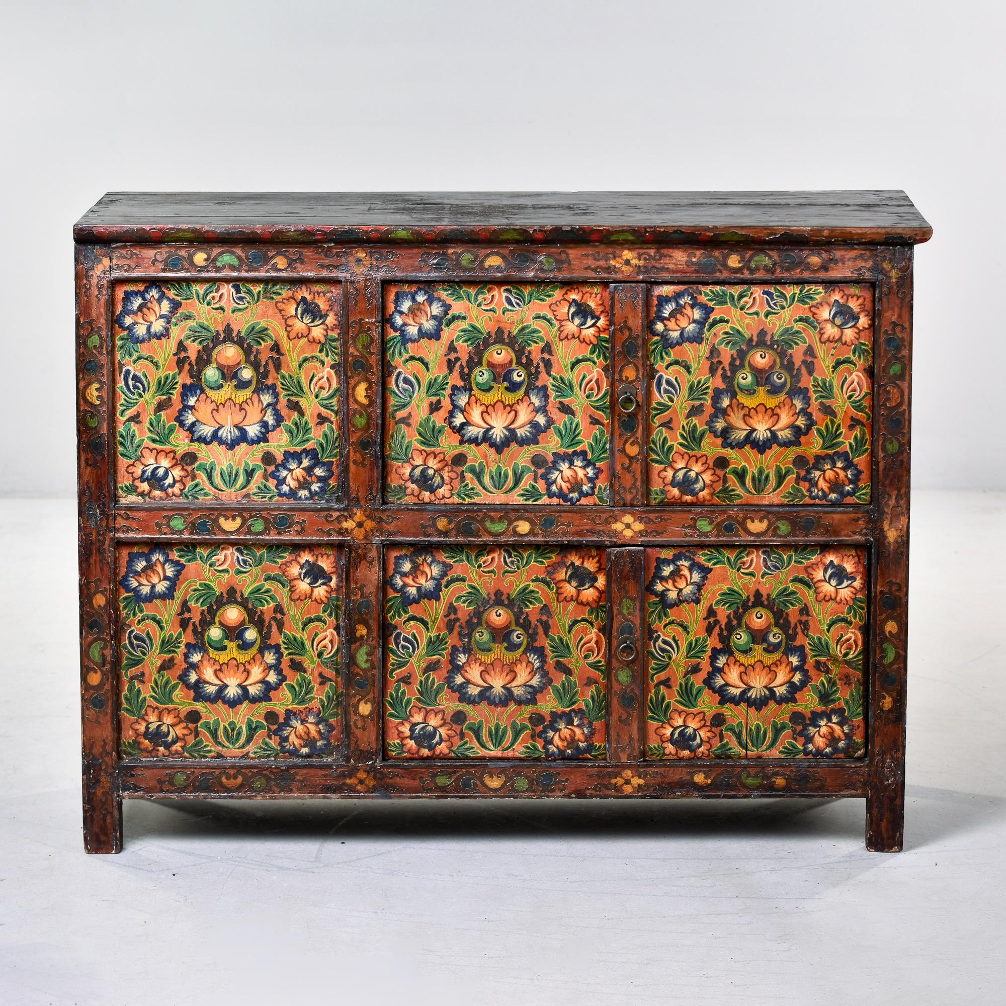 Circa 1910 Tibetan cabinet with vibrant hand painted finish depicting elaborate lotus blossom design across six panels on the front. Dominant color hues are blue, golds, greens, cream and a dusky persimmon. Cabinet opens at the front on two levels