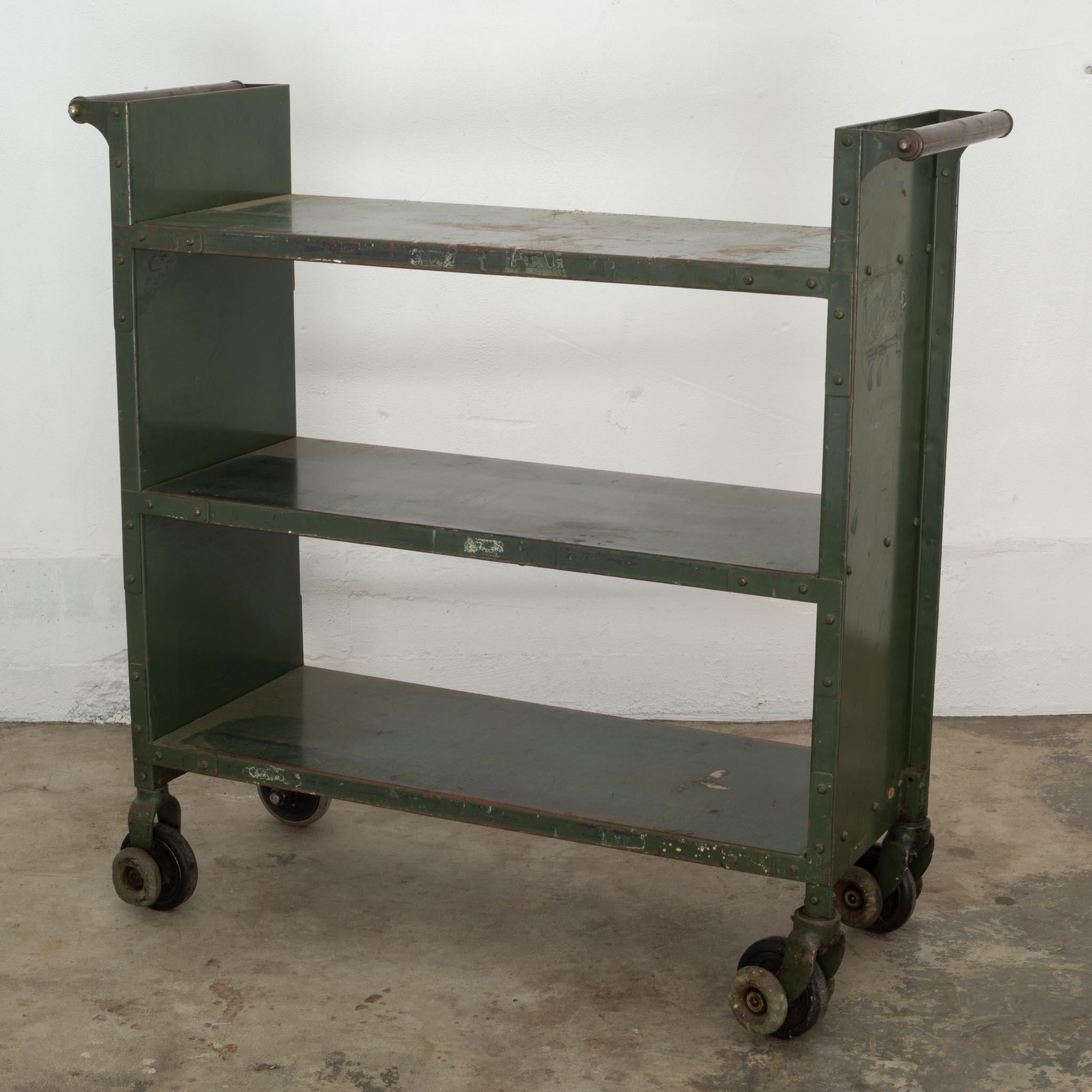 About

An industrial metal rolling library cart with two steel handles and multiple sets of rubber wheels on each foot. The wheels fit on a tracks that ran between the library stacks to reshelve books simpler, faster and quieter. The wheels on one