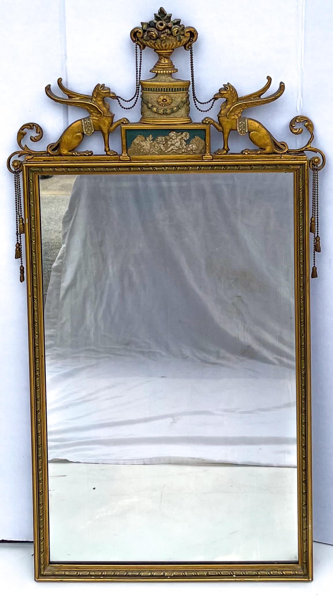 This is an early 20th century Italian mirror with floral pediment, carved griffins and draping brass tassels. There is a cherubic plaque that shows wear, and there is some wear throughout the gilt. It is unmarked.