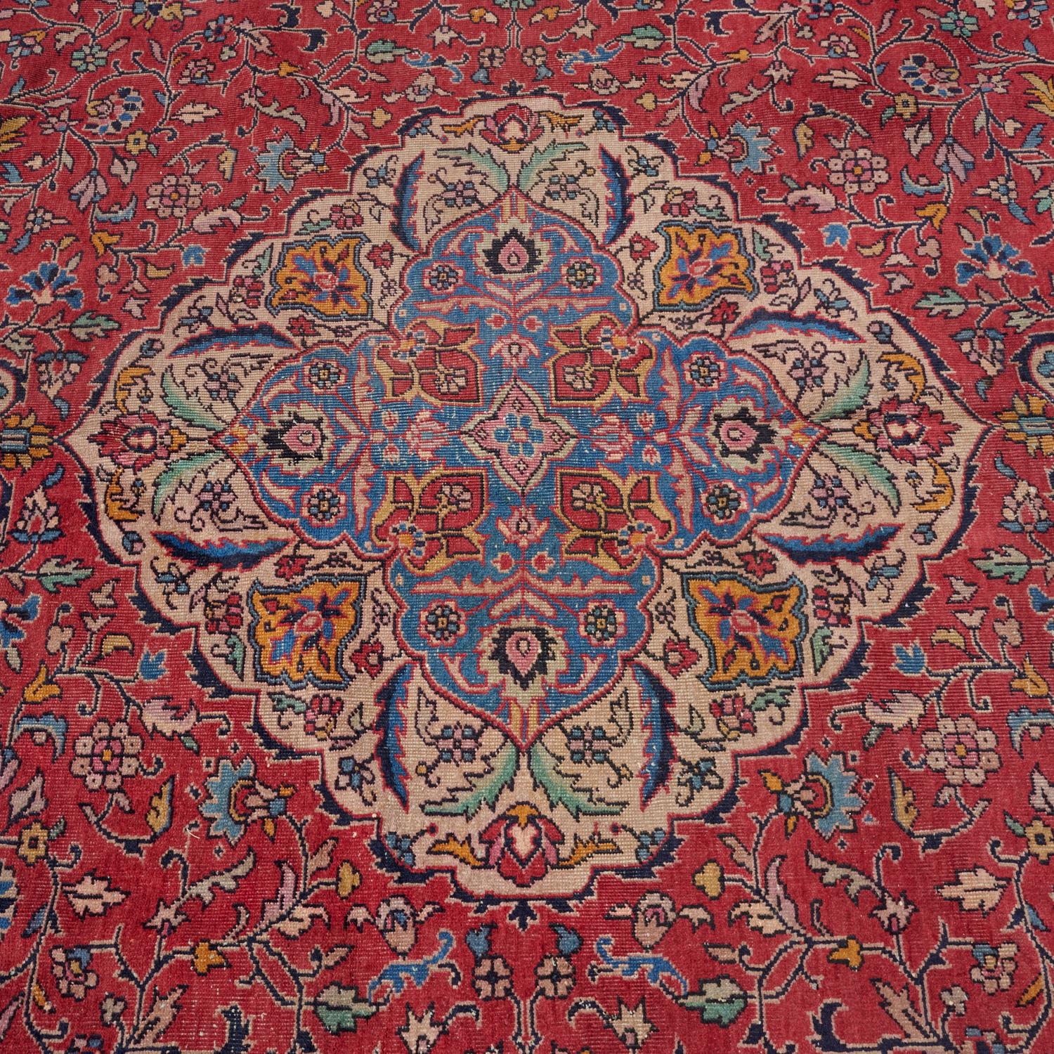 From the first half of the 20th c., Persia, a low pile Kashan room sized wool carpet. A traditional lozenge Kashan medallion is encompassed with an allover floral pattern on a red ground. Graceful corner spandrels flow into the red field.