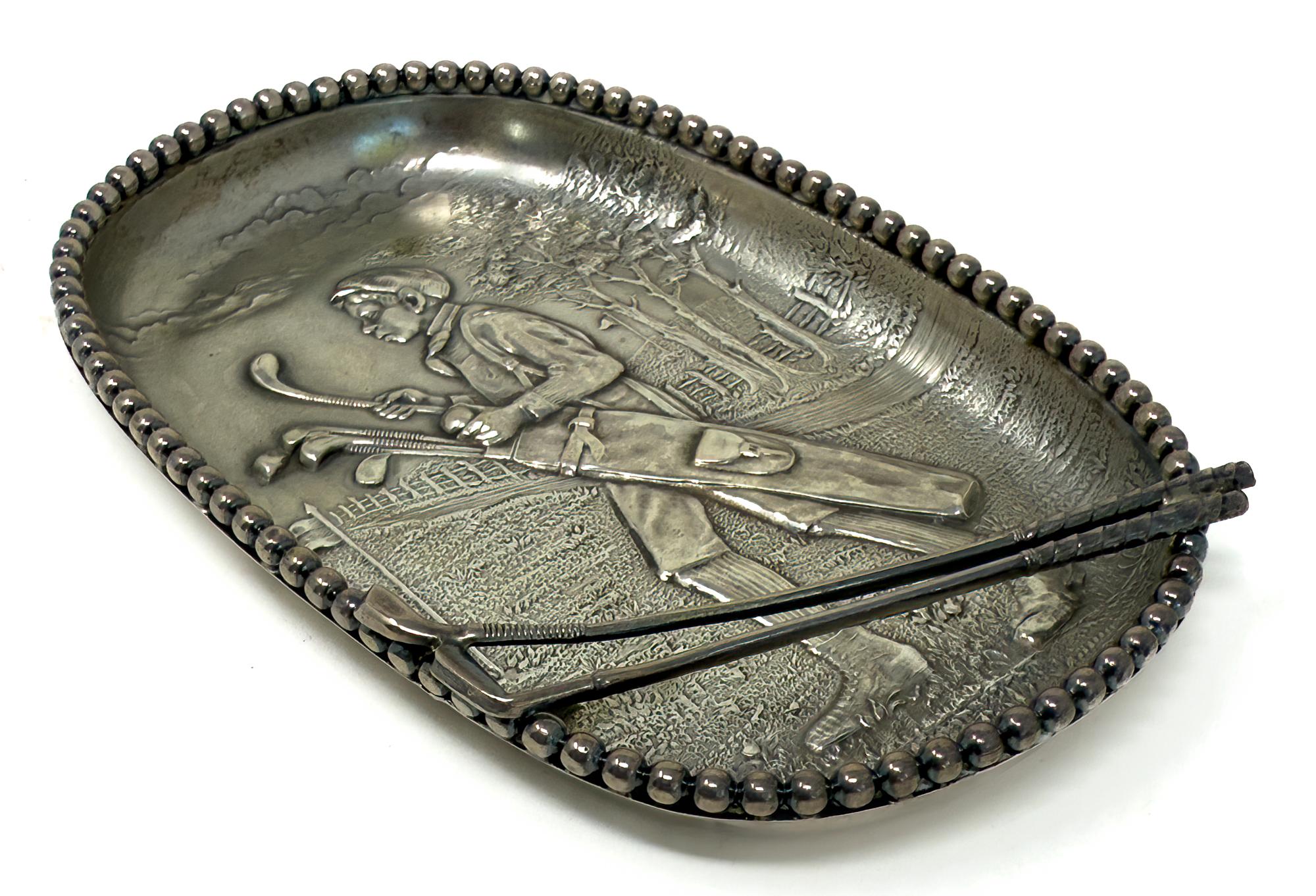 Un posacenere per sigari in argento sterling a tema golfistico di Unger Bros. dei primi anni del XX secolo. New Jersey. 
Un caddy afroamericano con una borsa sul campo da golf. Due mazze fissate nella parte inferiore del vassoio per raccogliere la