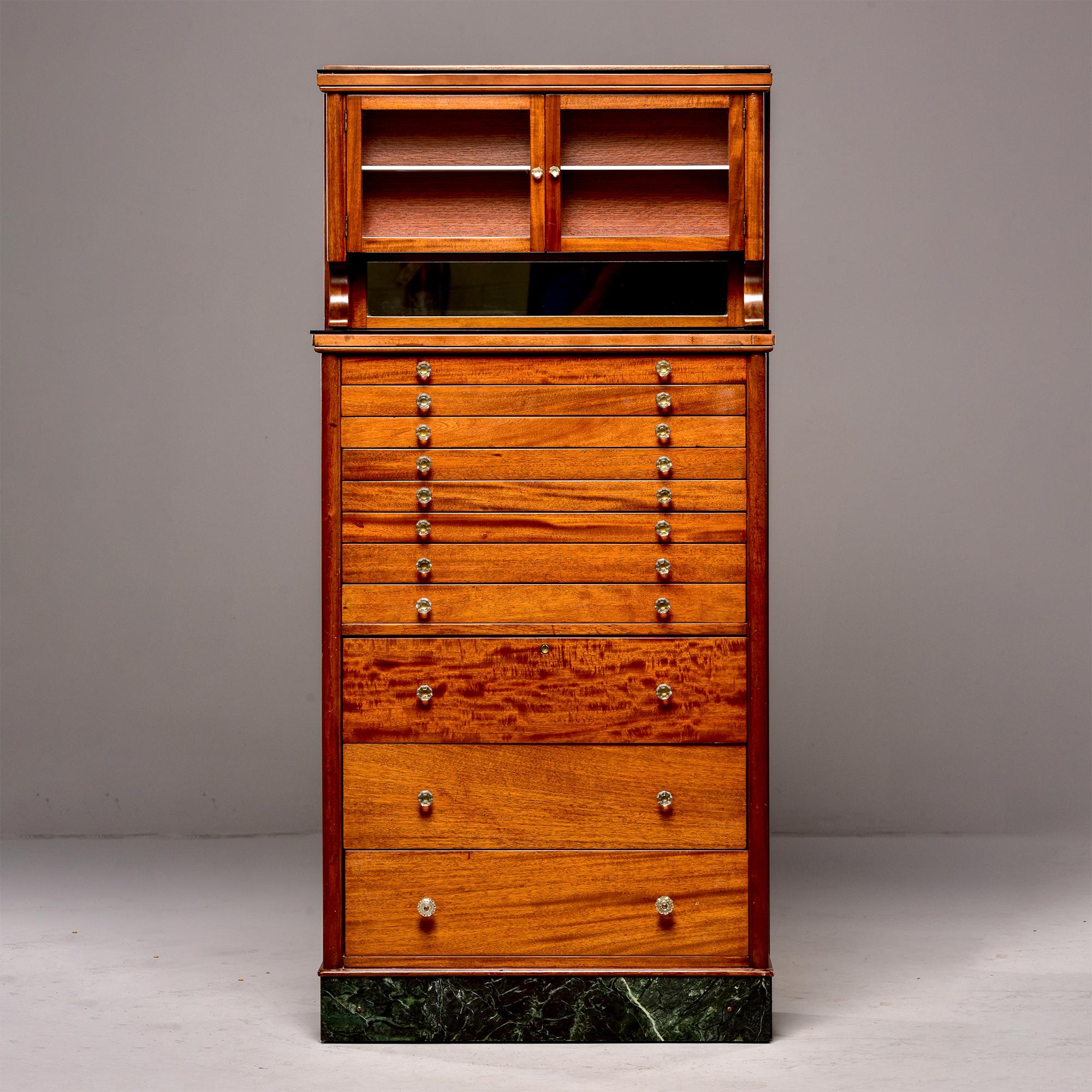 Circa 1920s American made medical / dental cabinet made of wood with base of 11 graduated sized drawers and a top section with glass front doors and internal shelf. Clear glass pulls/hardware. Unknown maker.