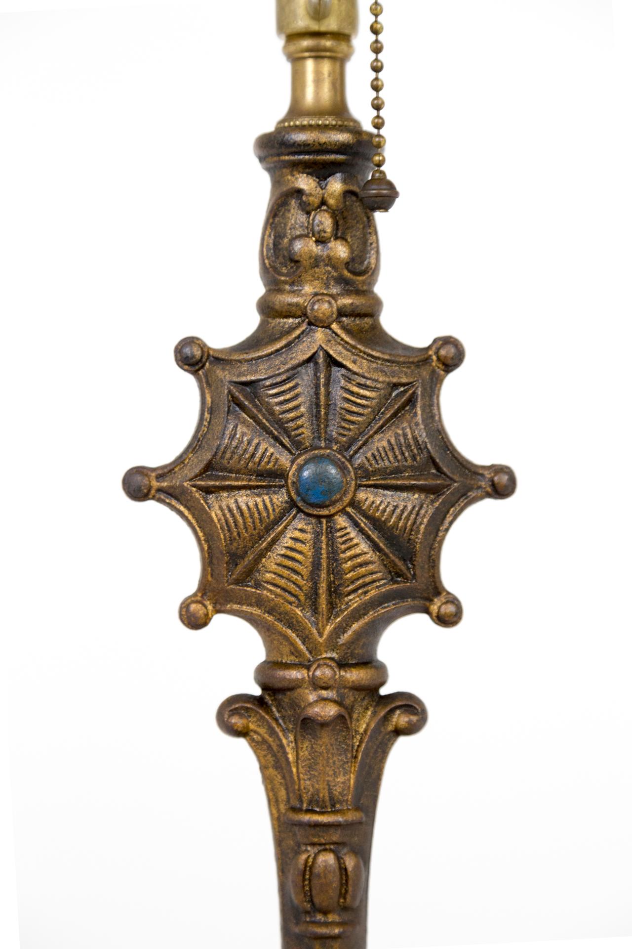 A brass table lamp with dark toned patina and gothic revival details; a radial spider web shape on the stem with a deep blue center dot, and C-curves on the base. With a pull chain, double, medium base, 3-way socket, original harp and finial; newly