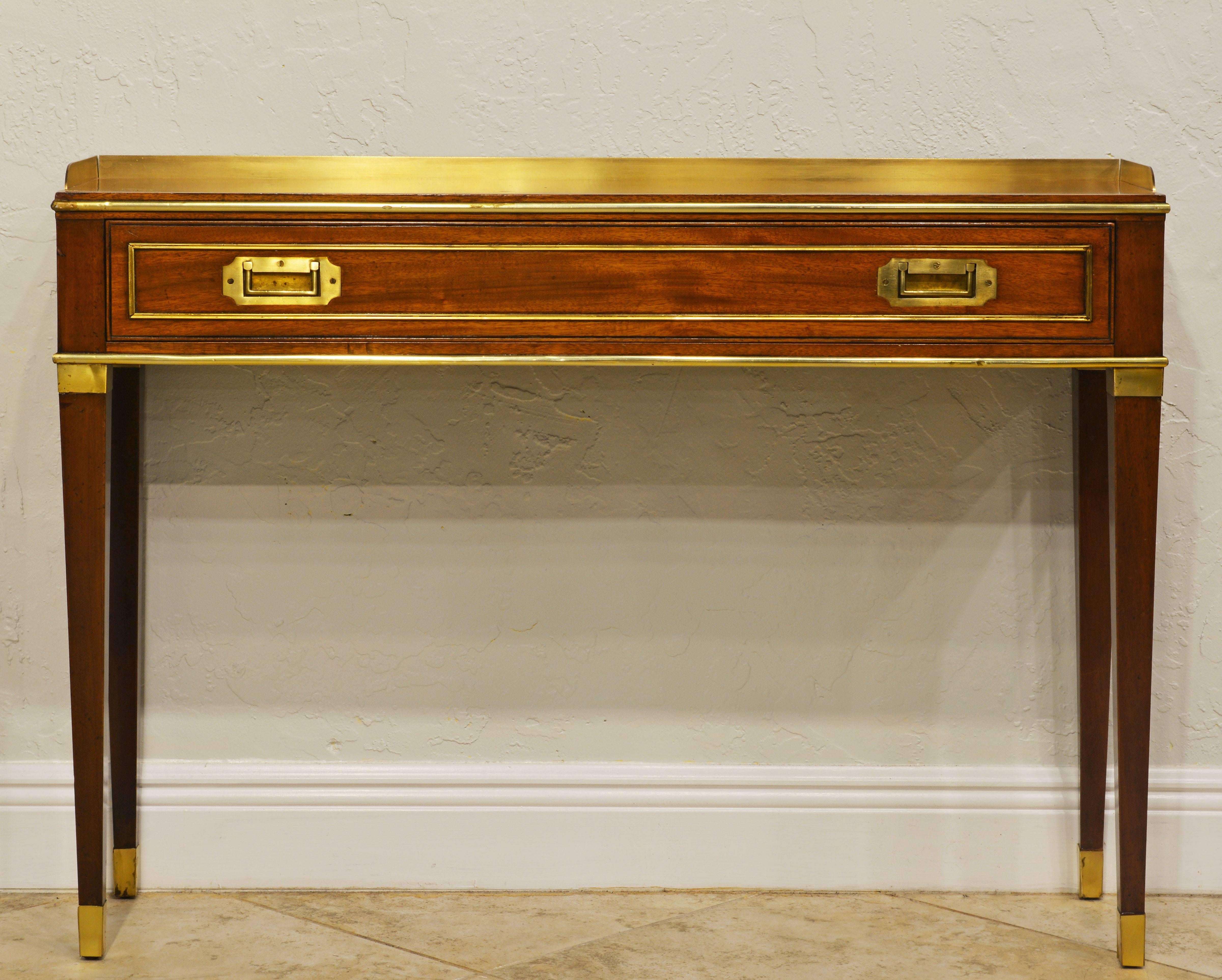 This fine and elegant console table features a brass edged mahogany top partly bordered by a brass gallery above a long drawer with recessed handles and brass trim resting on four square tapered legs surmounted by brass heads and ending in brass