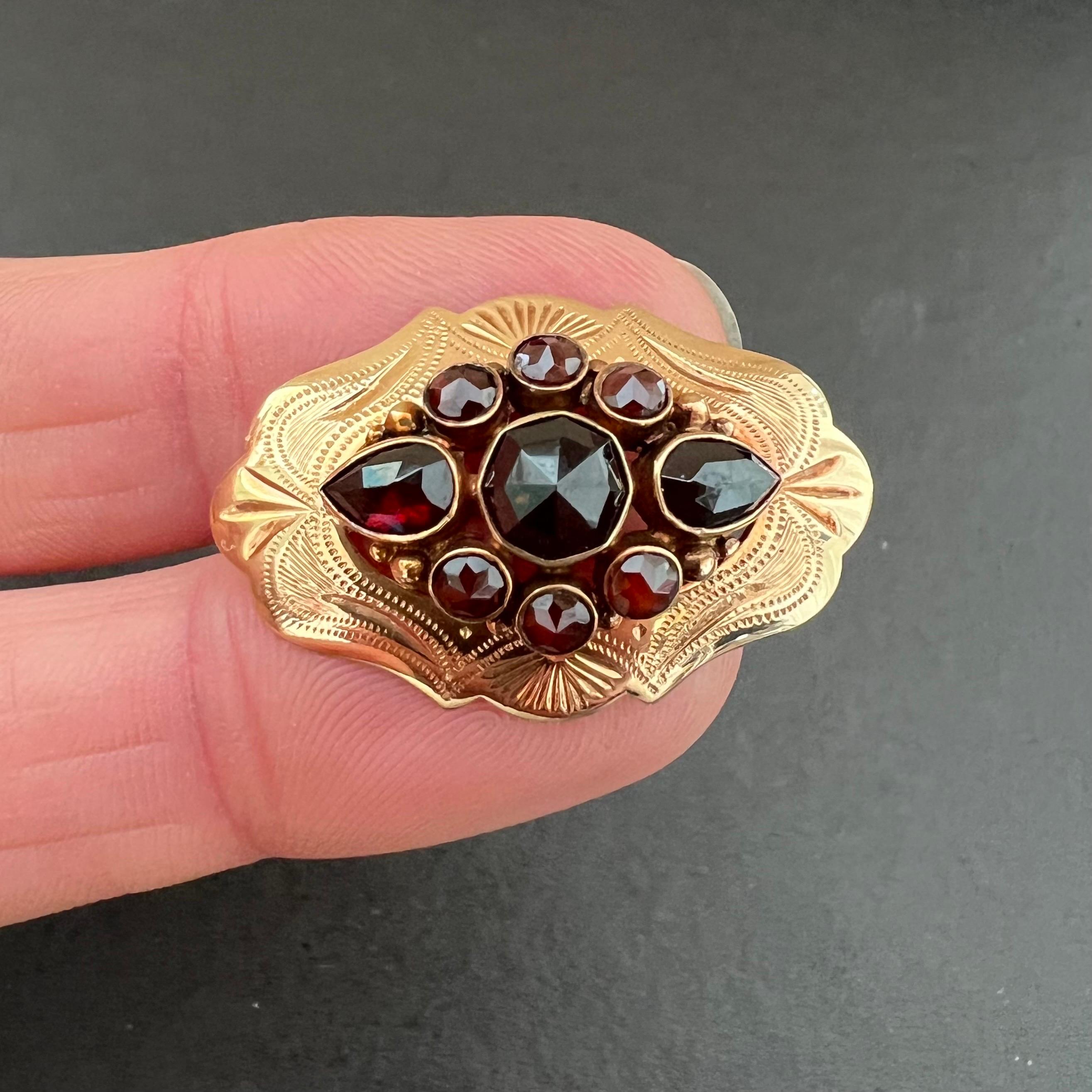 A 14 karat yellow gold oval brooch with an engraved scalloped edge. The brooch is set with a fantasy garnet entourage in the center with seven round and two drop-shaped faceted garnets. The garnets are rose cut and bezel set in the gold mounting.