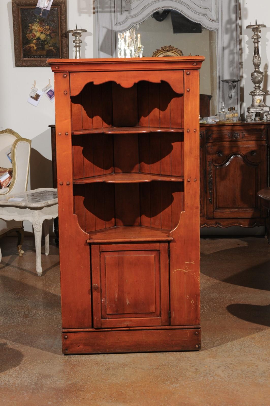 An American cherry corner cabinet from the early 20th century, with open shelves, single door and scalloped accents. Made in the USA during the early years of the 20th century, this corner cabinet features a beveled cornice sitting above three