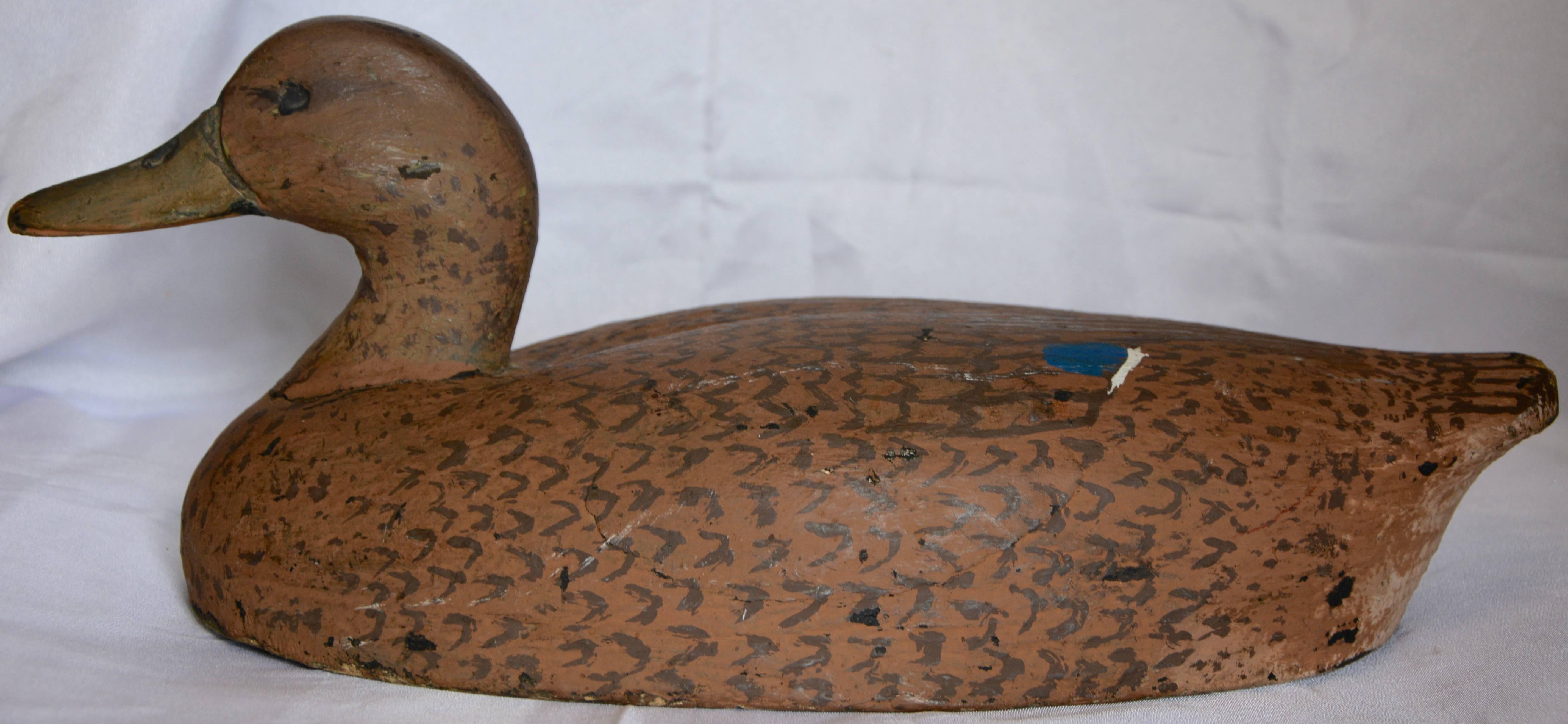 Featured is a hand-carved wooden duck decoy that has been creatively painted by the artist. He is a soft brown with darker brown details for his feathers. The wing tips are highlighted with a splash of blue and white.