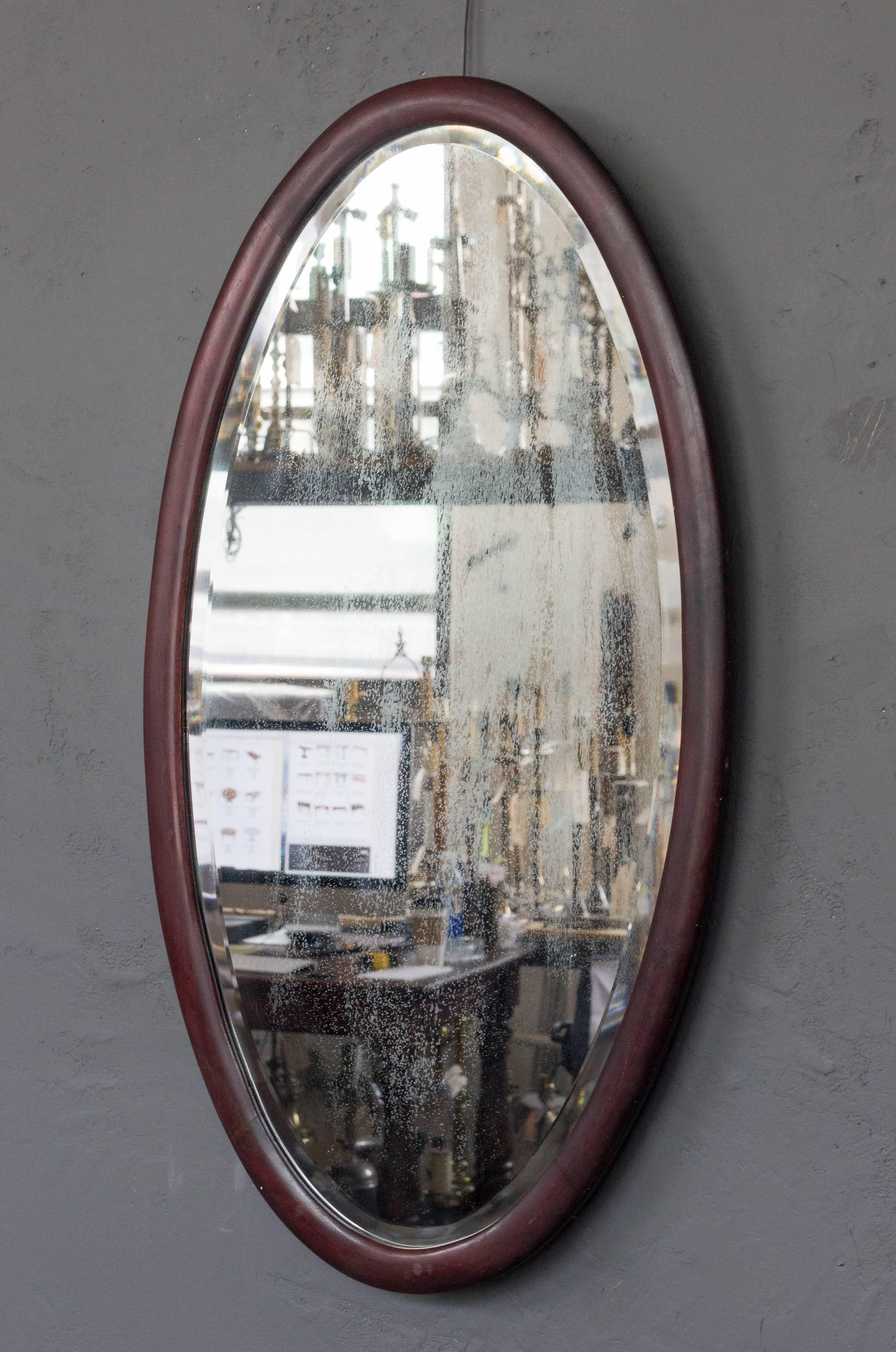 Oval shaped mirror with bevel in mahogany frame. Glass mirror is original and has an antique finish. This mirror had originally been attached a matching vanity/chest of drawers but has been modified to hang on the wall.