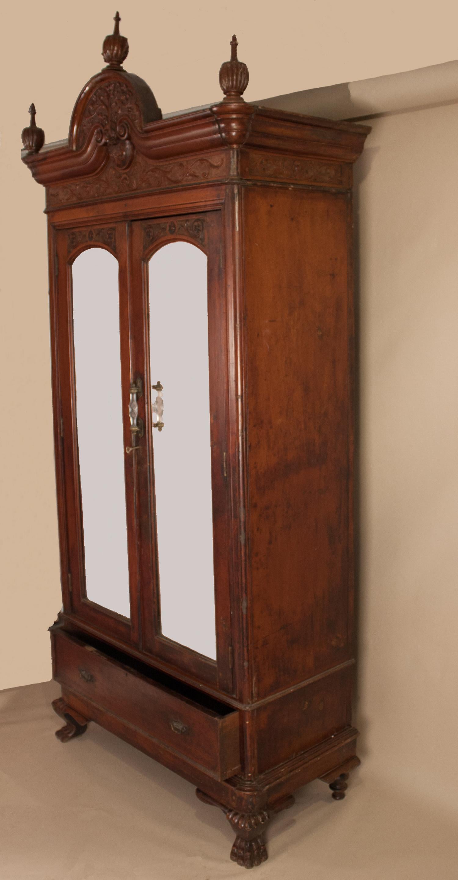 A tall and splendid piece, this Anglo-Indian carved mahogany armoire was constructed in three pieces for greater ease of transport. The wardrobe features a bevelled mirrored center section with lock and key, which opens to four shelves and two