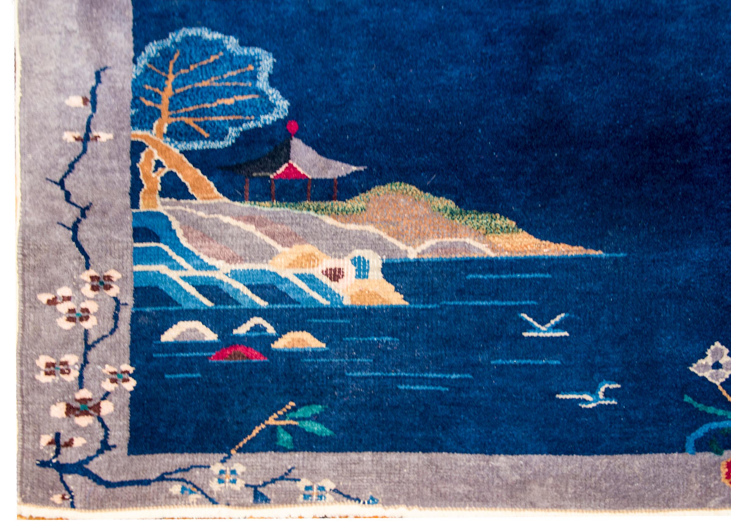An early 20th century Chinese Art Deco rug with a beautiful indigo field with a pagoda under a tree on an island surrounded by a grey border with blossoming peonies, chrysanthemum, and cherry blossoms.